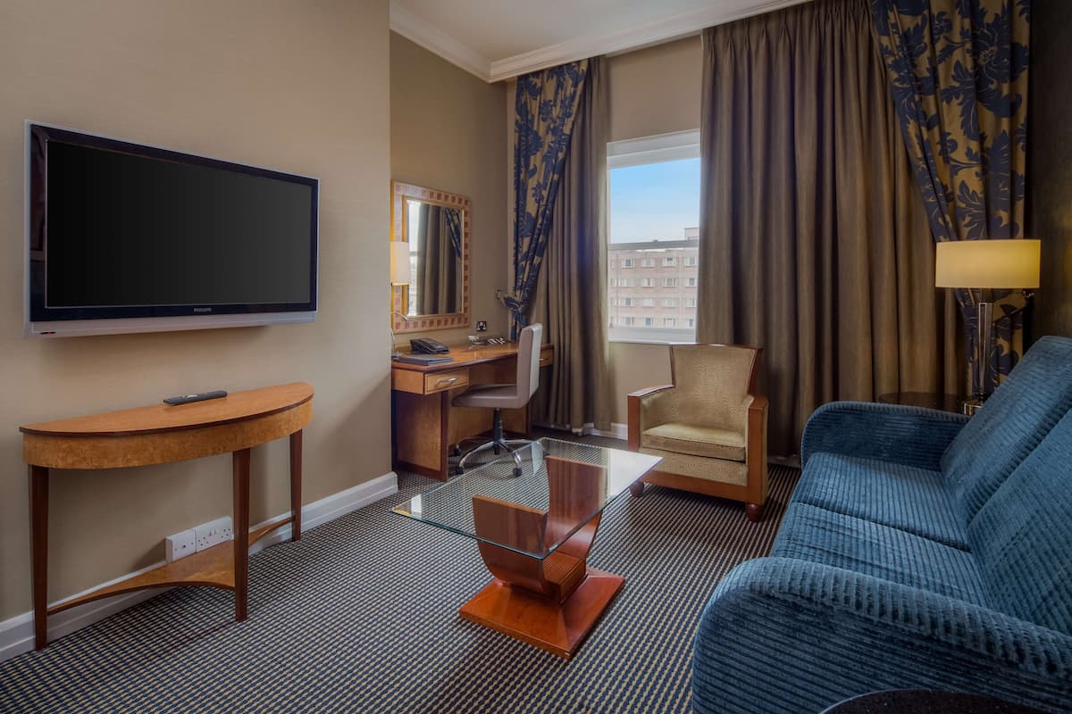 A hotel room with a flat-screen TV, a desk with a chair, a sofa, and a large window with curtains.