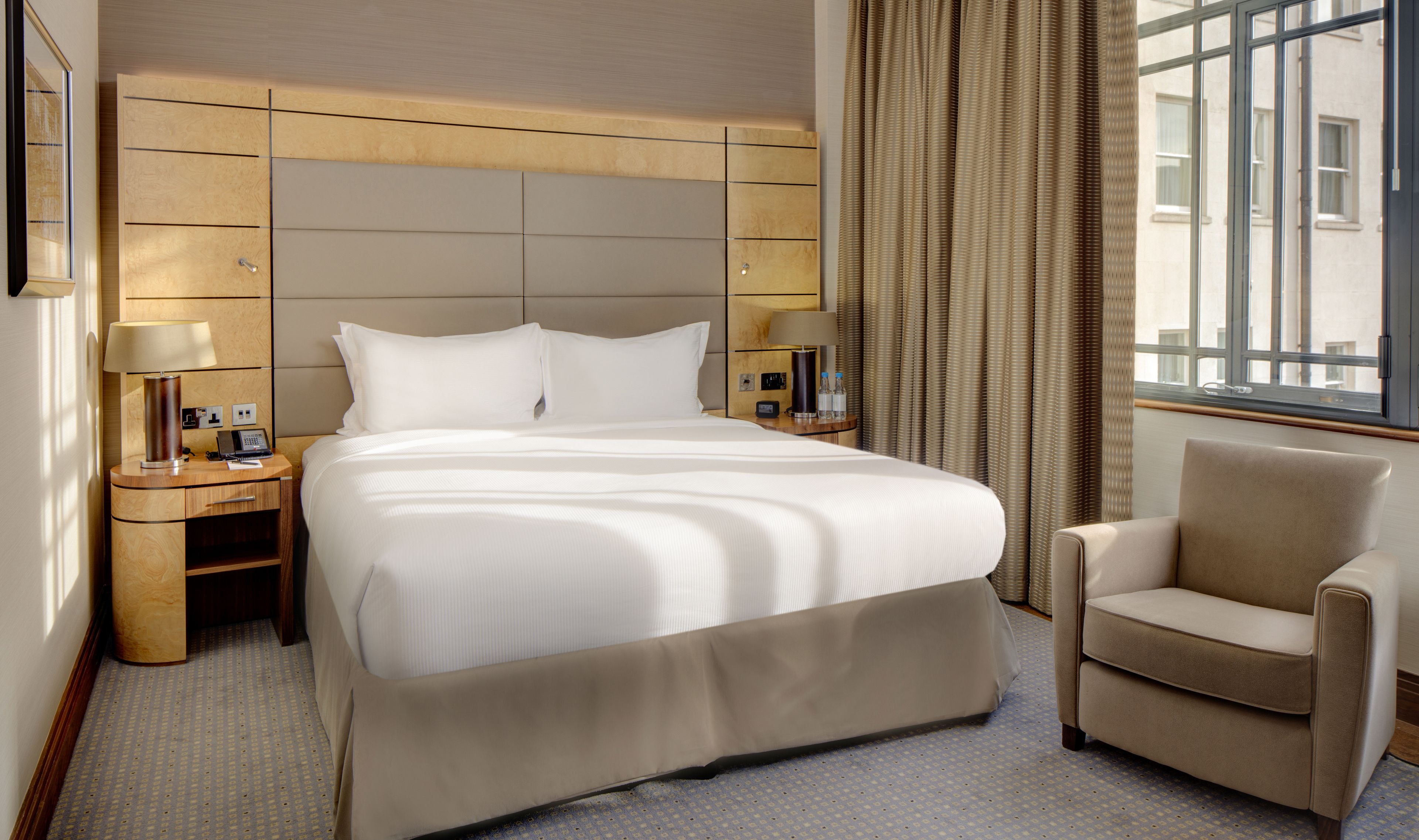A hotel room with a large bed, two bedside tables, a chair, and a window with curtains.