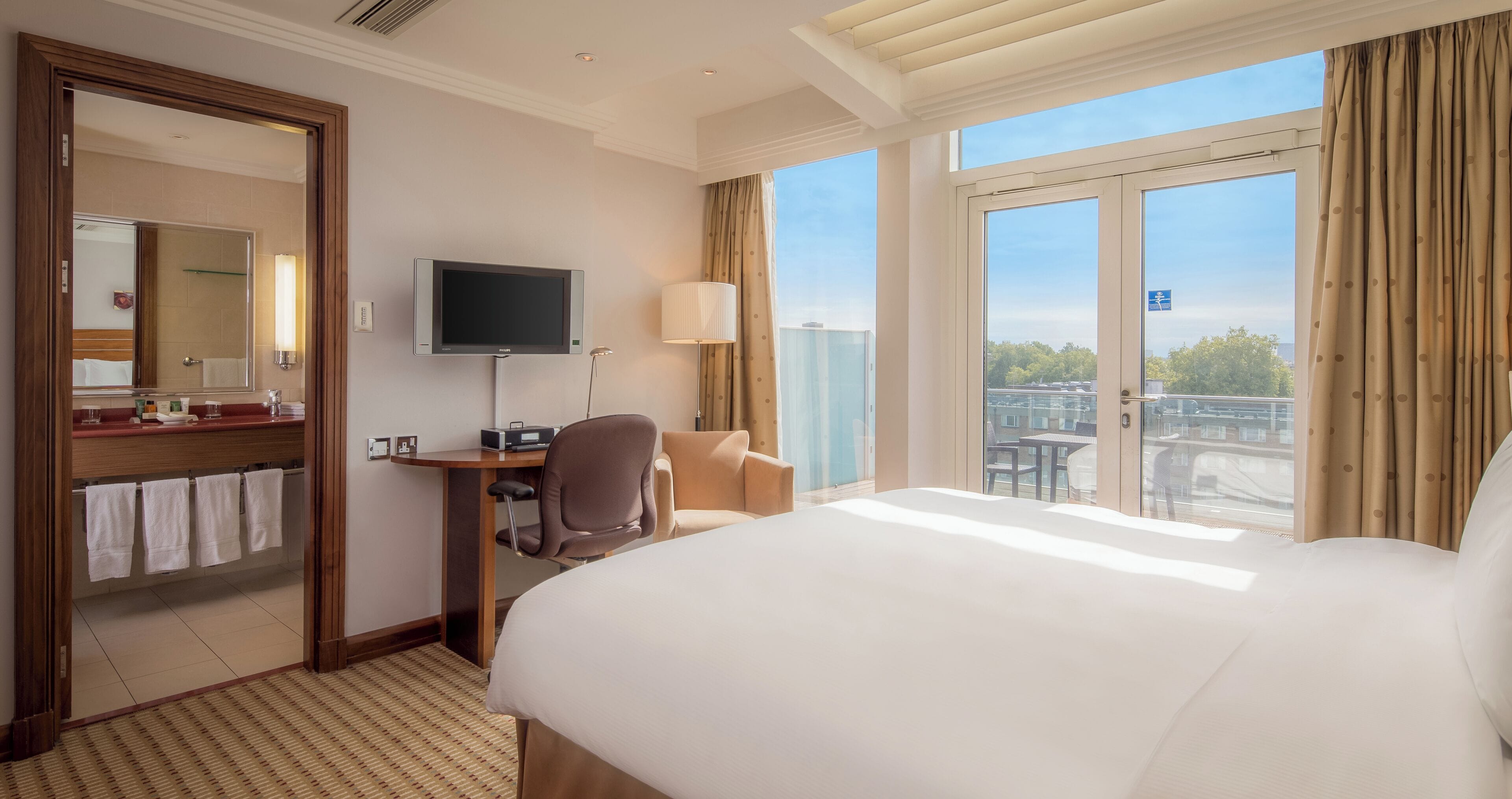 Habitación de hotel con una cama grande, un escritorio con silla, un televisor y vistas al exterior por una puerta corredera.