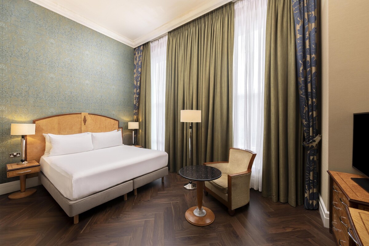 A hotel room with a bed, a chair, a table, a lamp, and a TV.