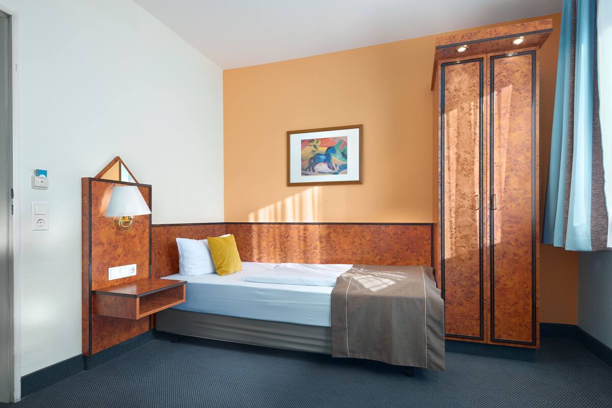 A hotel room with a bed, a nightstand, a lamp, a wardrobe, and a painting on the wall.