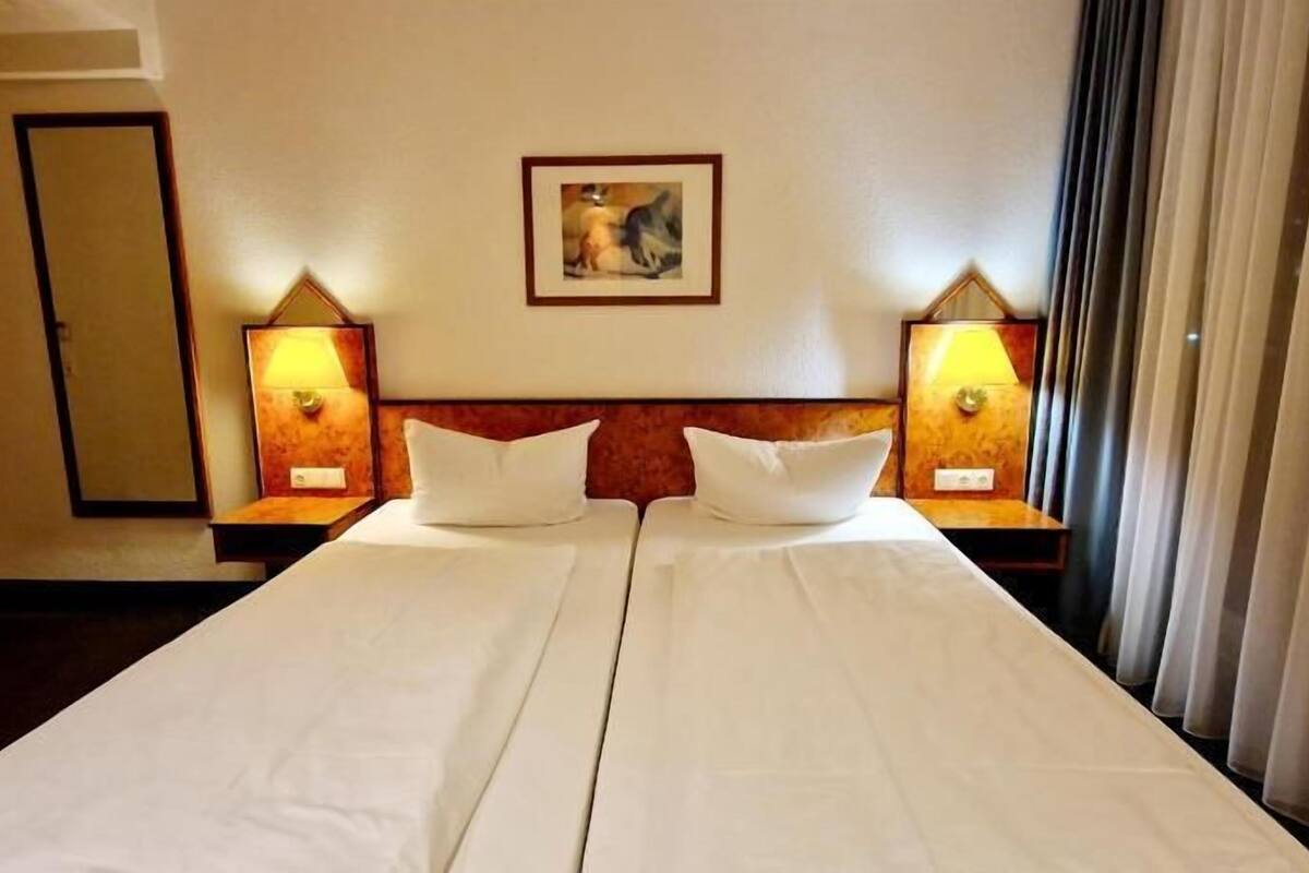 A hotel room with a large bed, two bedside lamps, a painting on the wall, and a window with curtains.