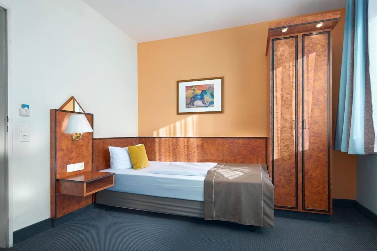 A hotel room with a bed, a nightstand, a lamp, a wardrobe, and a painting on the wall.