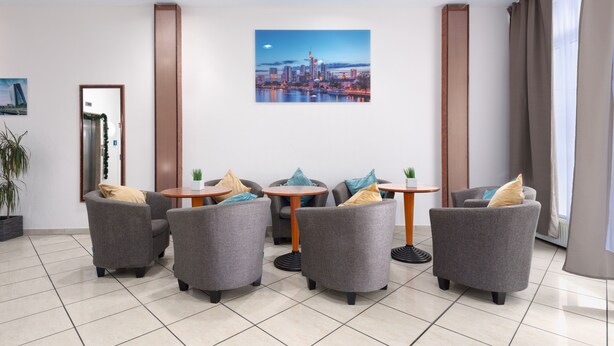 A modern lounge area with grey armchairs, round wooden tables, and a cityscape painting on the wall. - Trip Inn Hotel Minerva (Frankfurt)
