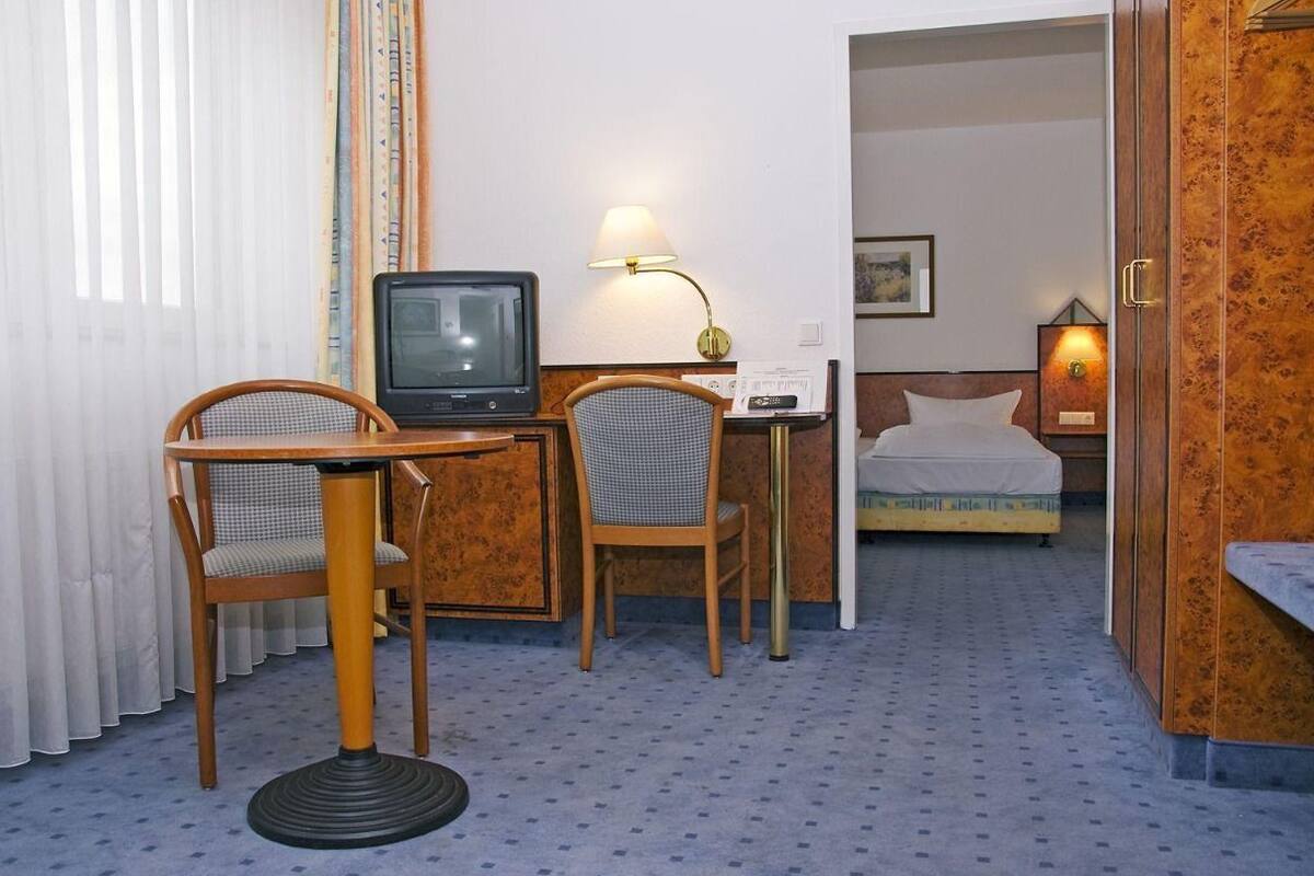 A hotel room with a bed, a desk with a television, two chairs, a lamp, and a door leading to another room.