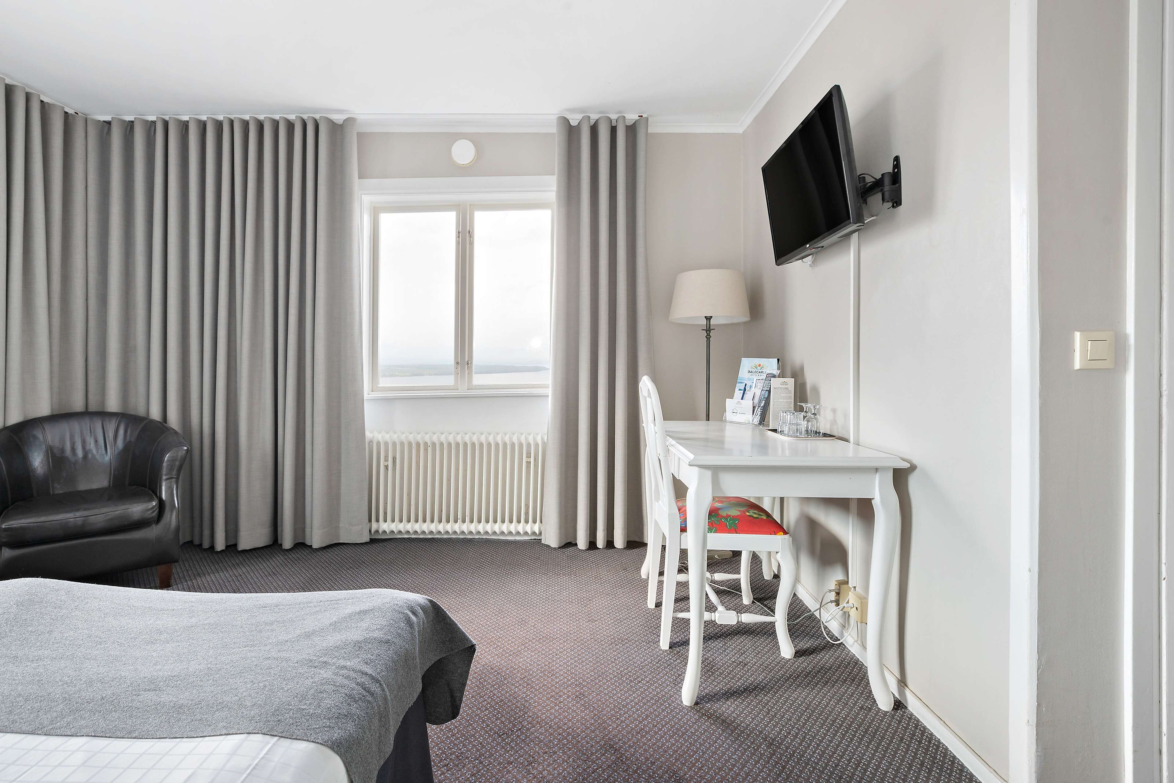 A hotel room with a bed, a desk with a chair, a television mounted on the wall, and a window with curtains.
