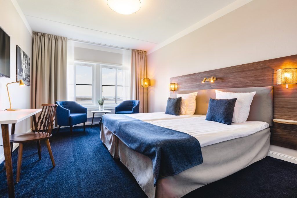 A hotel room with a large bed, a desk, two chairs, a window, and wall-mounted lights.