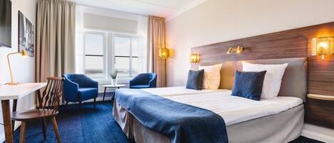 A hotel room with a large bed, a desk, two chairs, a window, and wall-mounted lights.