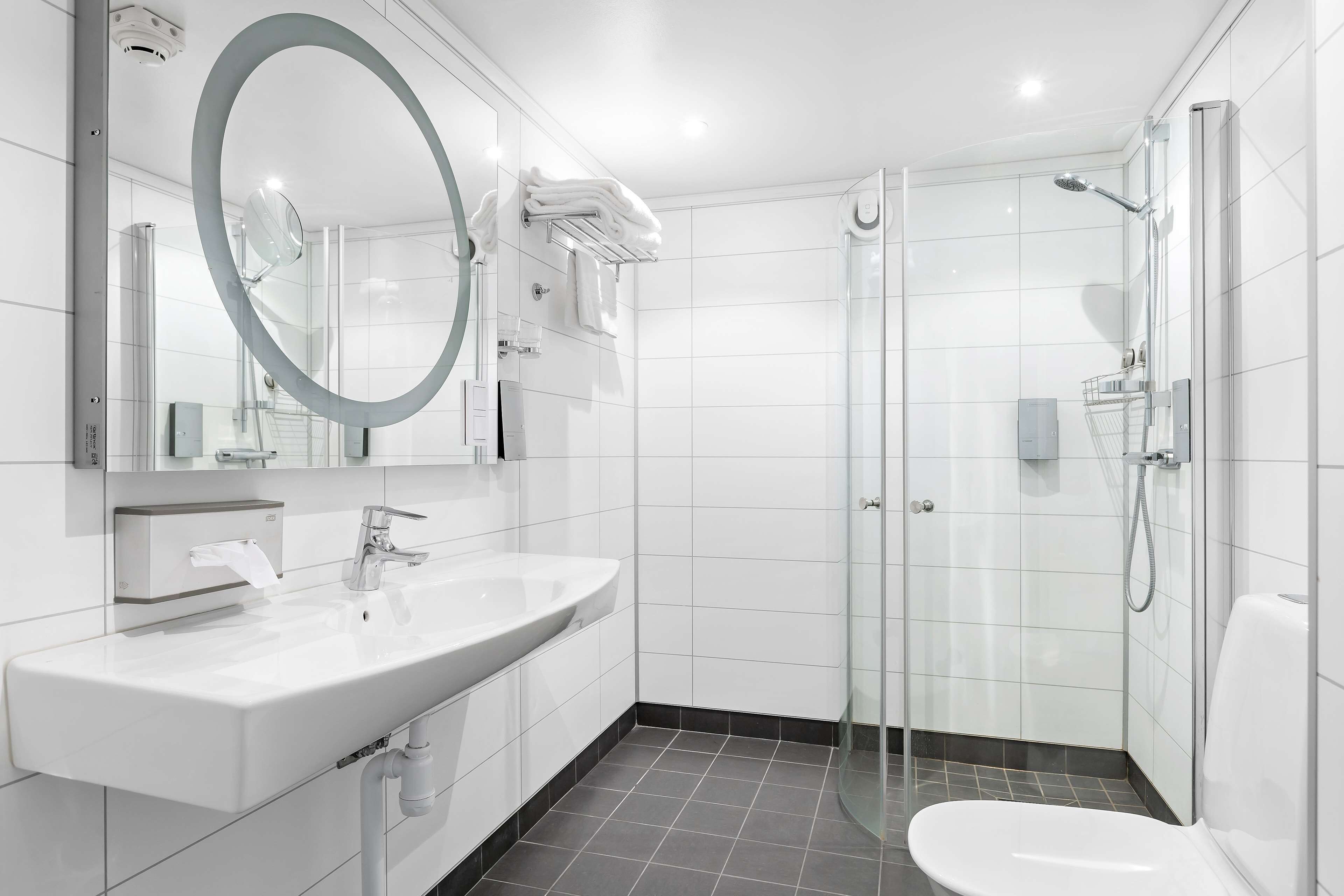 Un baño moderno con lavamanos, espejo y retrete blancos, y una zona de ducha con paredes de vidrio.