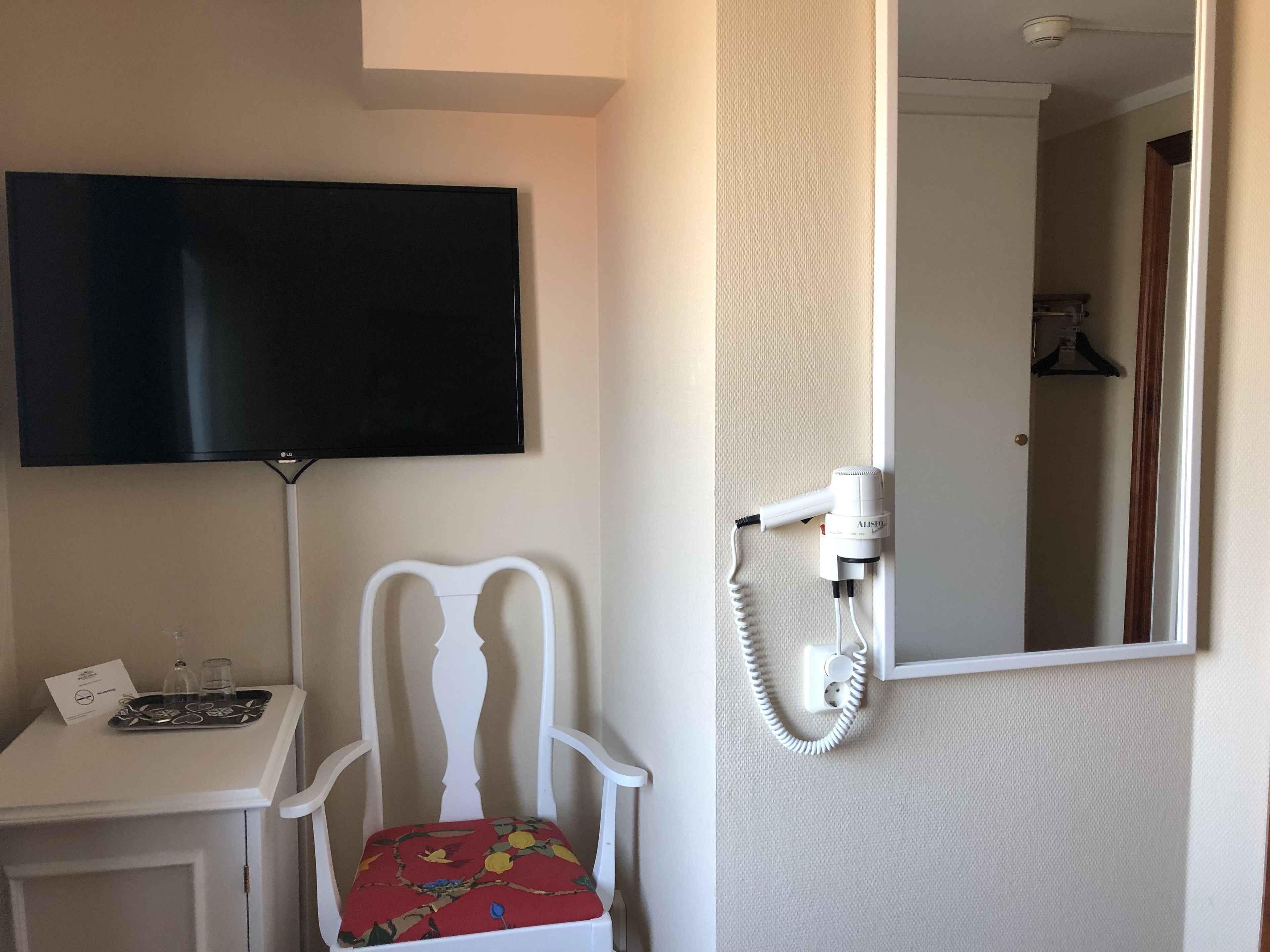 Habitación de hotel con televisor de pantalla plana montado en la pared, una silla blanca con cojín estampado, una mesita con una bandeja de vasos y un secador de pelo conectado a un enchufe.