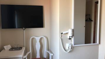 Habitación de hotel con televisor de pantalla plana montado en la pared, una silla blanca con cojín estampado, una mesita con una bandeja de vasos y un secador de pelo conectado a un enchufe.