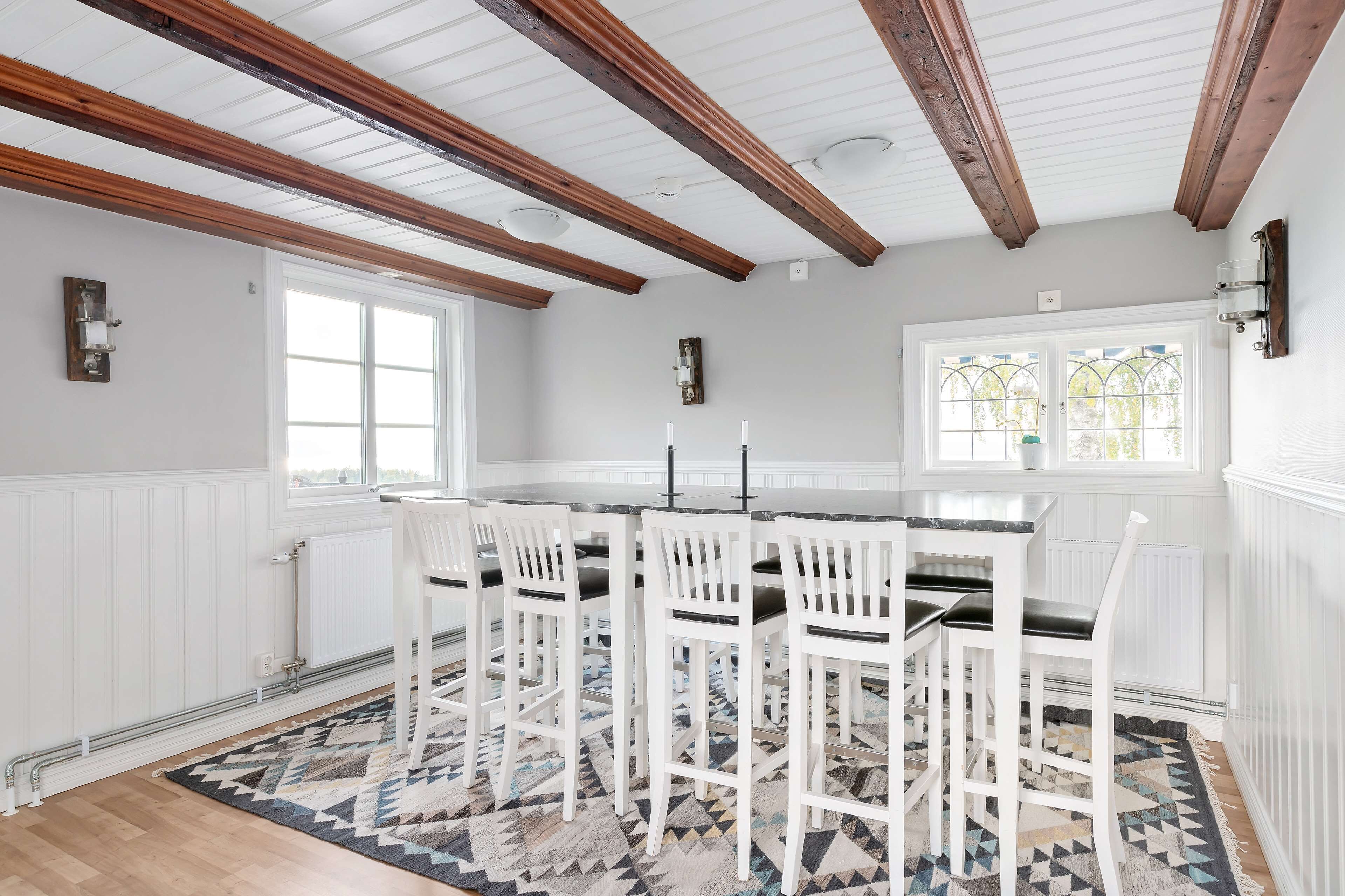 A kitchen with white cabinetry, a central island with bar stools, wooden beams on the ceiling, and a patterned rug.
