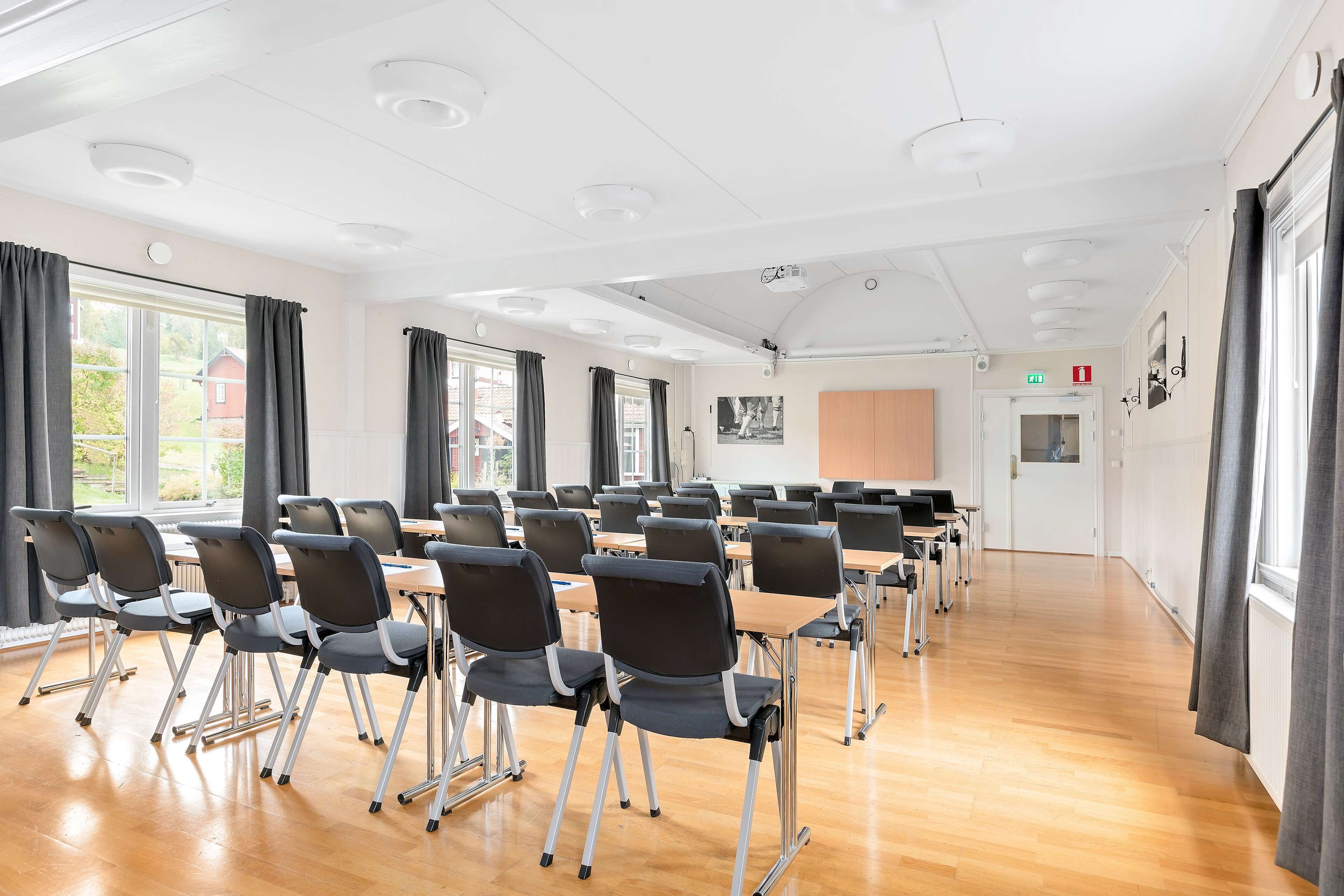 Konferenssihuone, jossa on riveissä tuoleja ja pöytiä, suuret ikkunat, joissa on verhot, ja valkoinen katto, jossa on upotetut valaisimet.