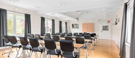 A conference room with rows of chairs and tables, large windows with curtains, and a white ceiling with recessed lighting.