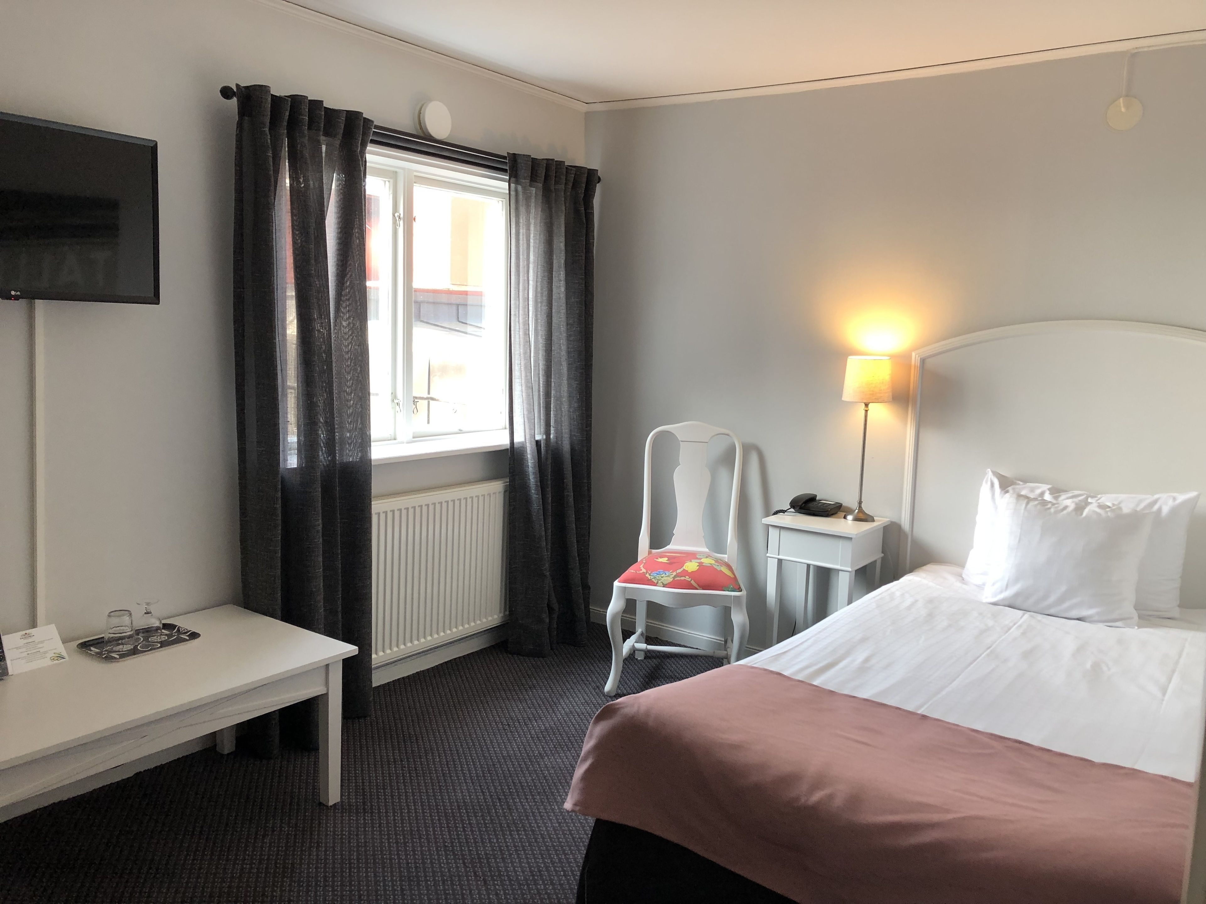 A hotel room with a bed, a desk, a chair, a lamp, and a TV.