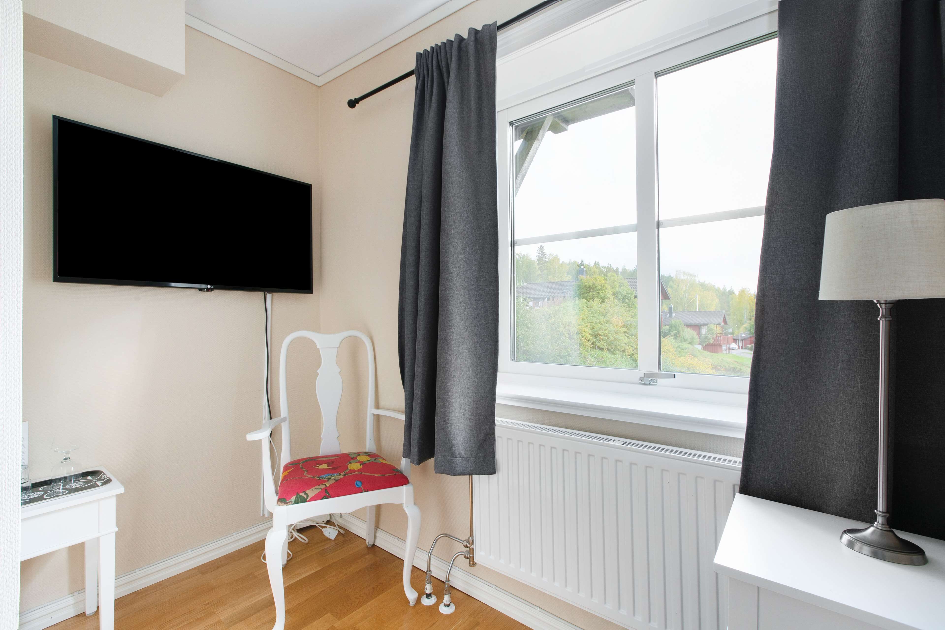 Une pièce avec un téléviseur fixé au mur, une chaise avec un coussin coloré, une fenêtre avec des rideaux et une lampe sur une table.