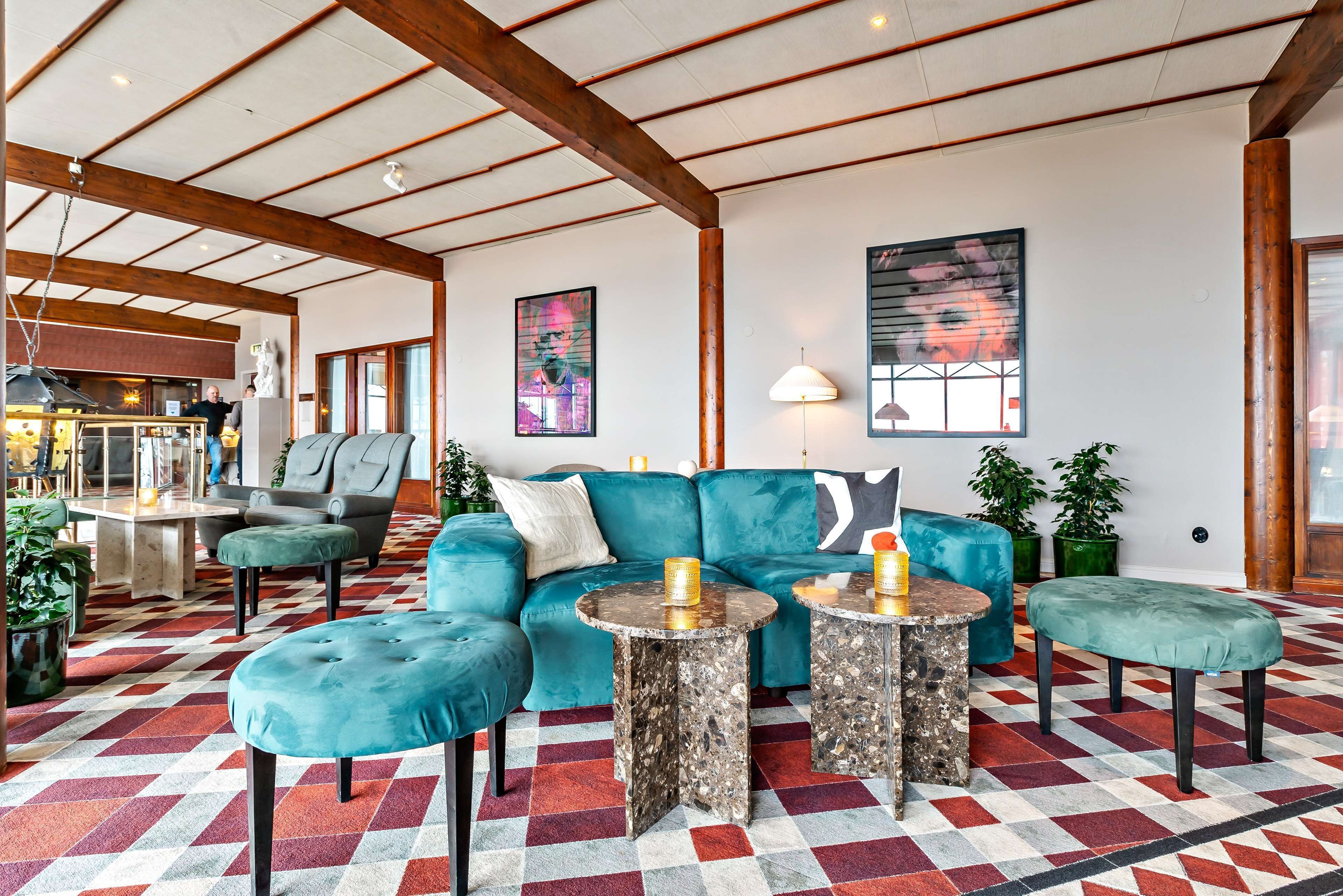 Een ruime kamer met een hoge plafon, een blauwgroene bank, ronde tafels en een kamerbreed tapijt met een patroon.