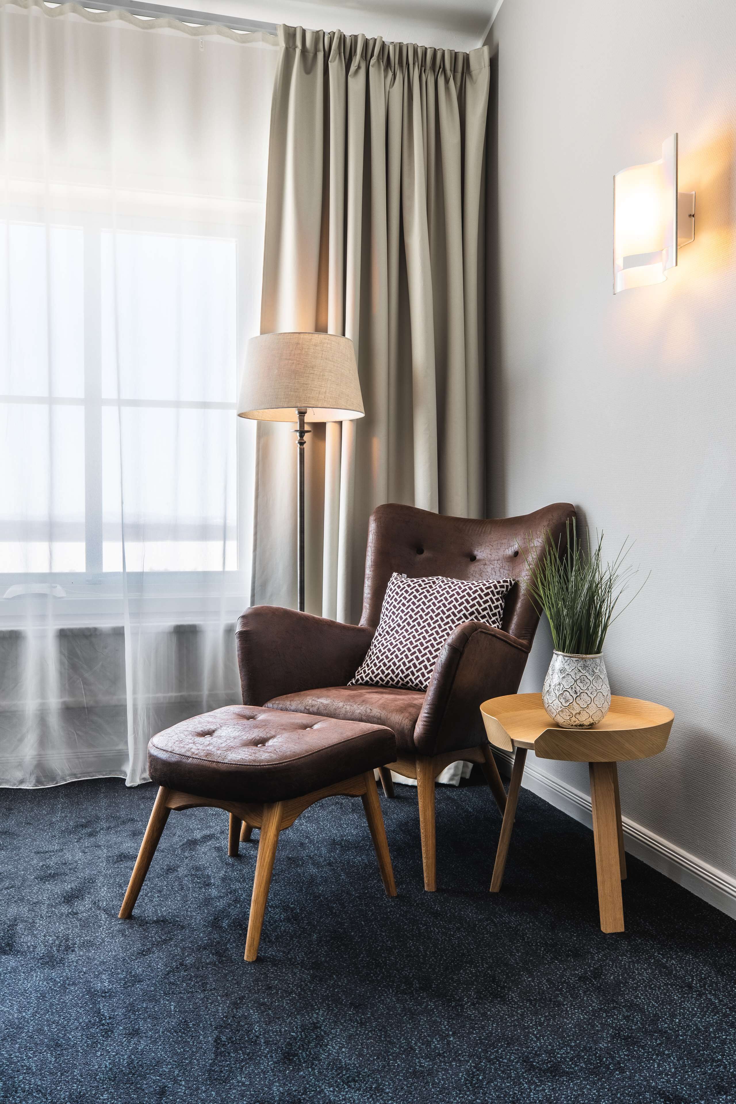 A room with a brown leather armchair, a small round table with a potted plant, a floor lamp, and a window with sheer curtains.