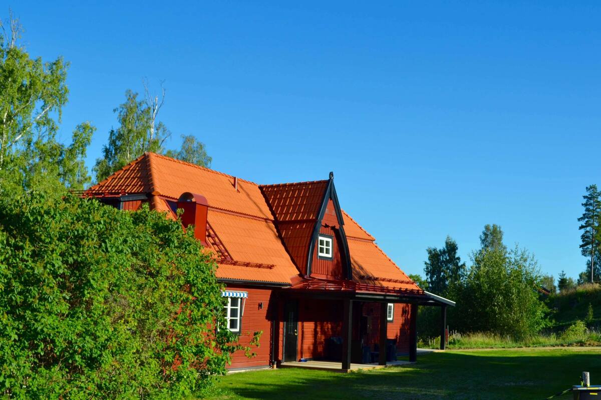 A red wooden house with a sloped roof, surrounded by greenery and trees.
