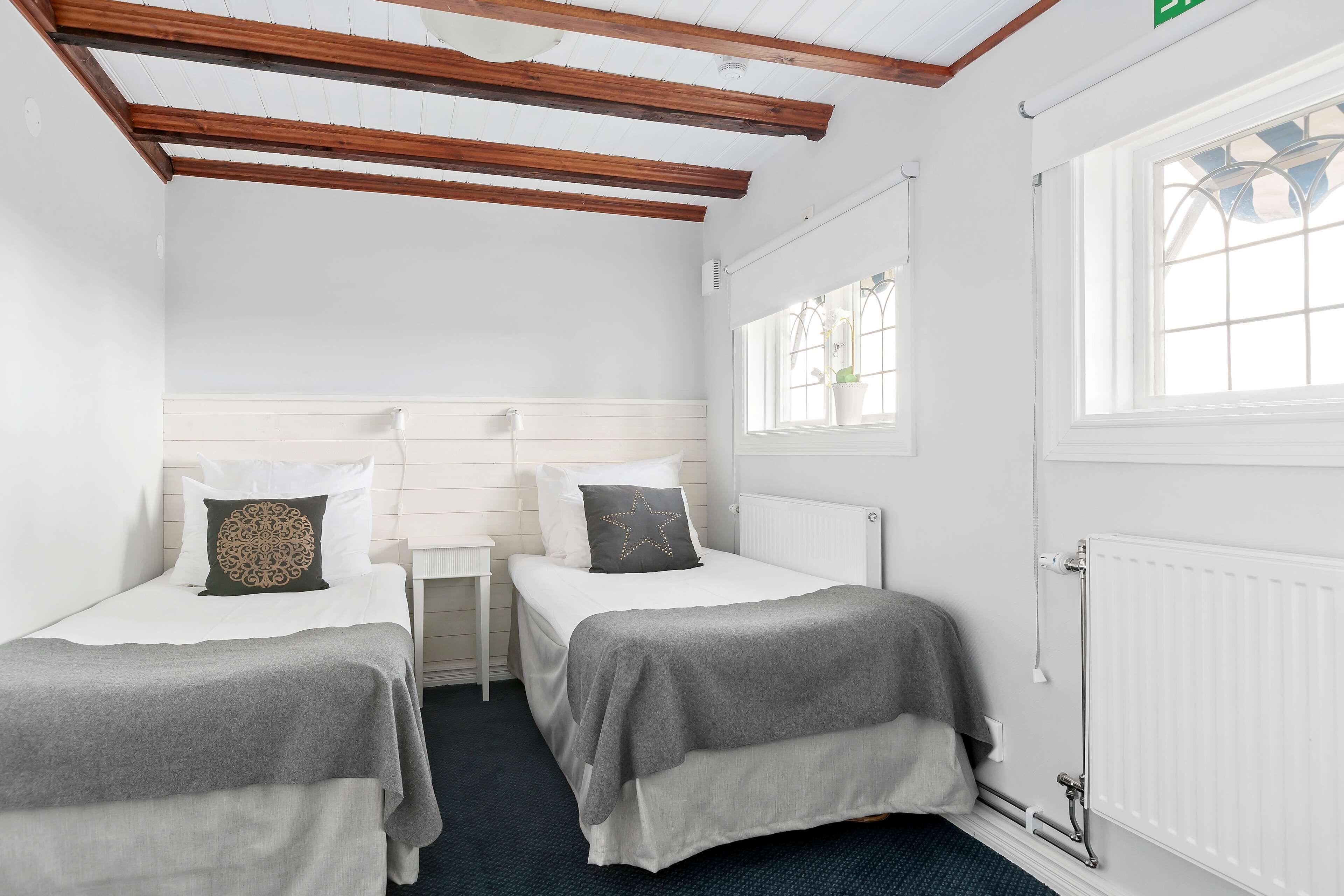 Ein Zimmer mit zwei Einzelbetten, weißen Wänden, Holzbalken an der Decke, einem Fenster mit Aussicht und einer Heizung.