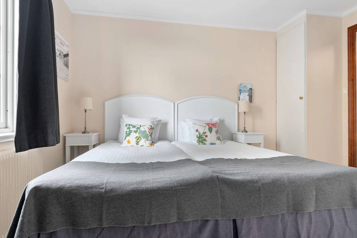 A double bed with a grey bedspread and floral pillows, two bedside tables with lamps, a window with a black curtain, and a door to the right.