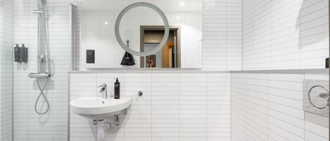 Une salle de bain moderne avec un mur recouvert de carreaux blancs, un lavabo avec un miroir, une douchette à main et une poubelle.