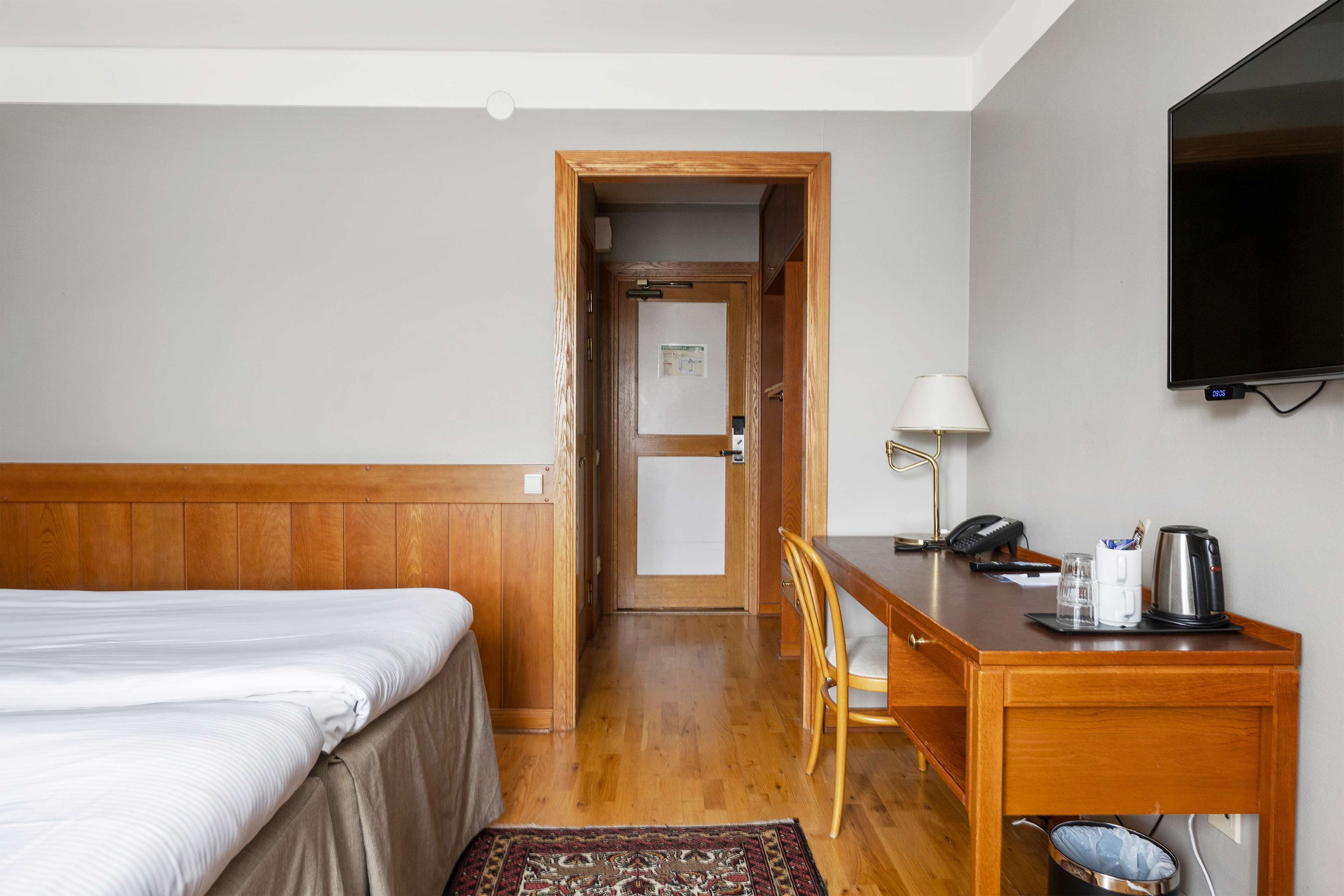 Habitación de hotel con una cama de madera, un escritorio con lámpara, un televisor y un pequeño cubo de basura.