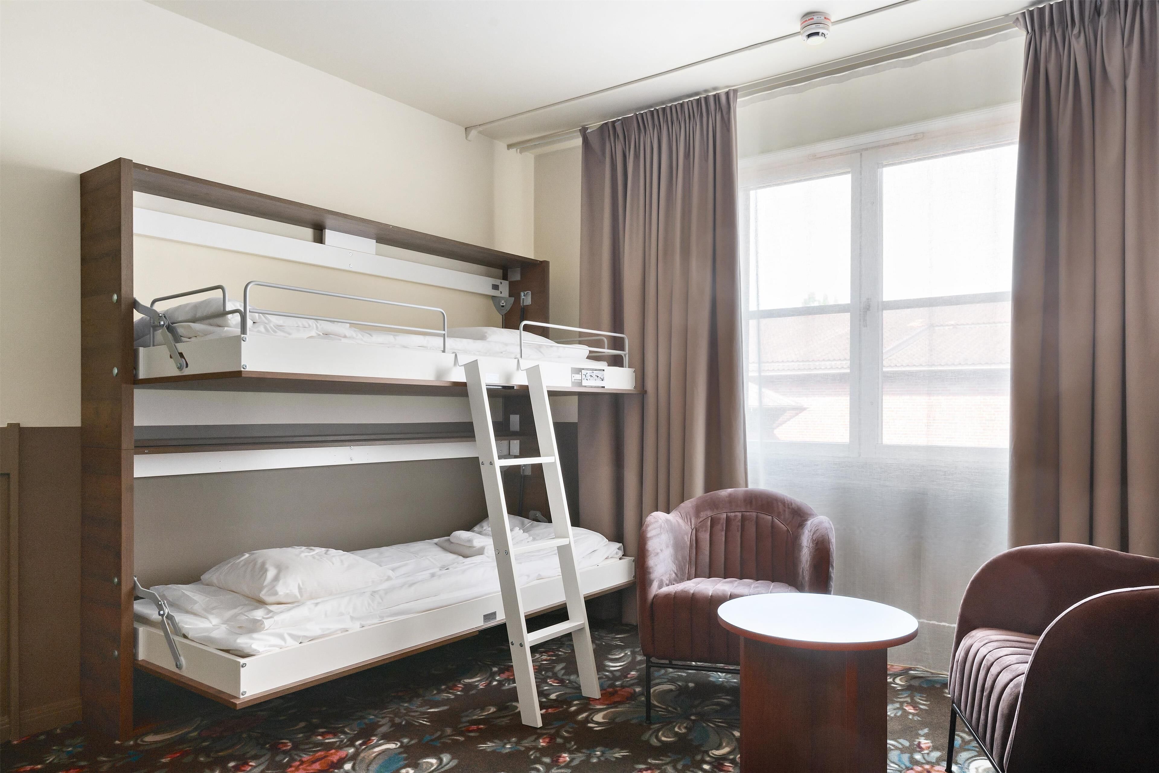 Una cama individual con escalera, una silla marrón, una mesita redonda y una ventana con cortinas.