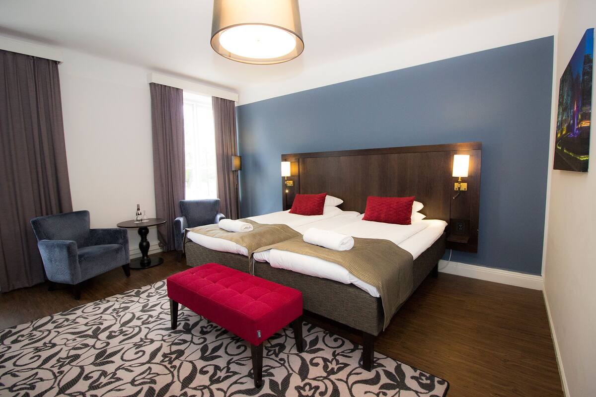 A hotel room with a large bed, a red bench, a small table, and a window with curtains.