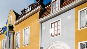 A building with a yellow facade and a grey section, featuring a traditional architectural style with a prominent window and a decorative entrance. - Grand Hotel Alingsås (Alingsas)