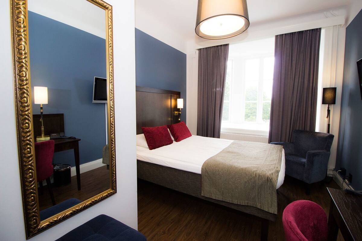 A hotel room with a bed, a desk, a chair, a mirror, and a window with curtains.