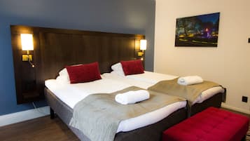 Quarto de hotel com duas camas, cada uma com travesseiro vermelho, roupa de cama branca e manta cinza. Há um banco vermelho e um quadro na parede.