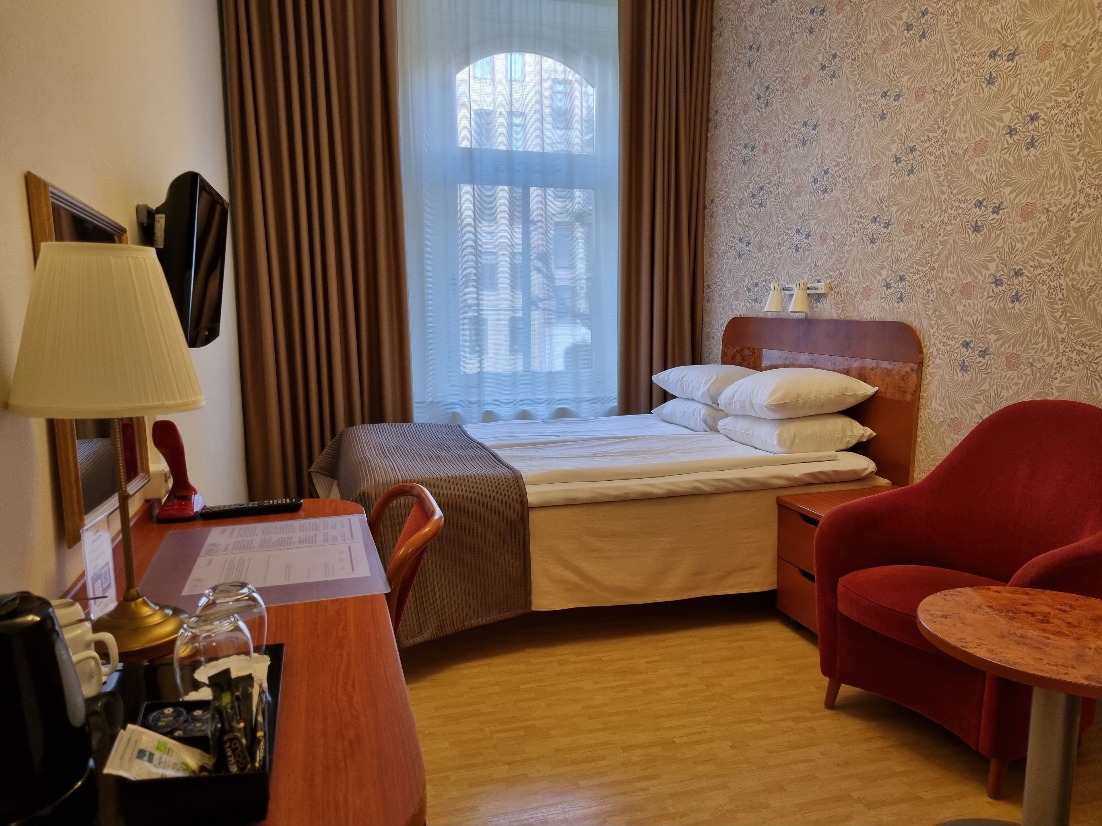 A hotel room with a bed, a desk with a lamp, a chair, a small table, and a window with curtains.