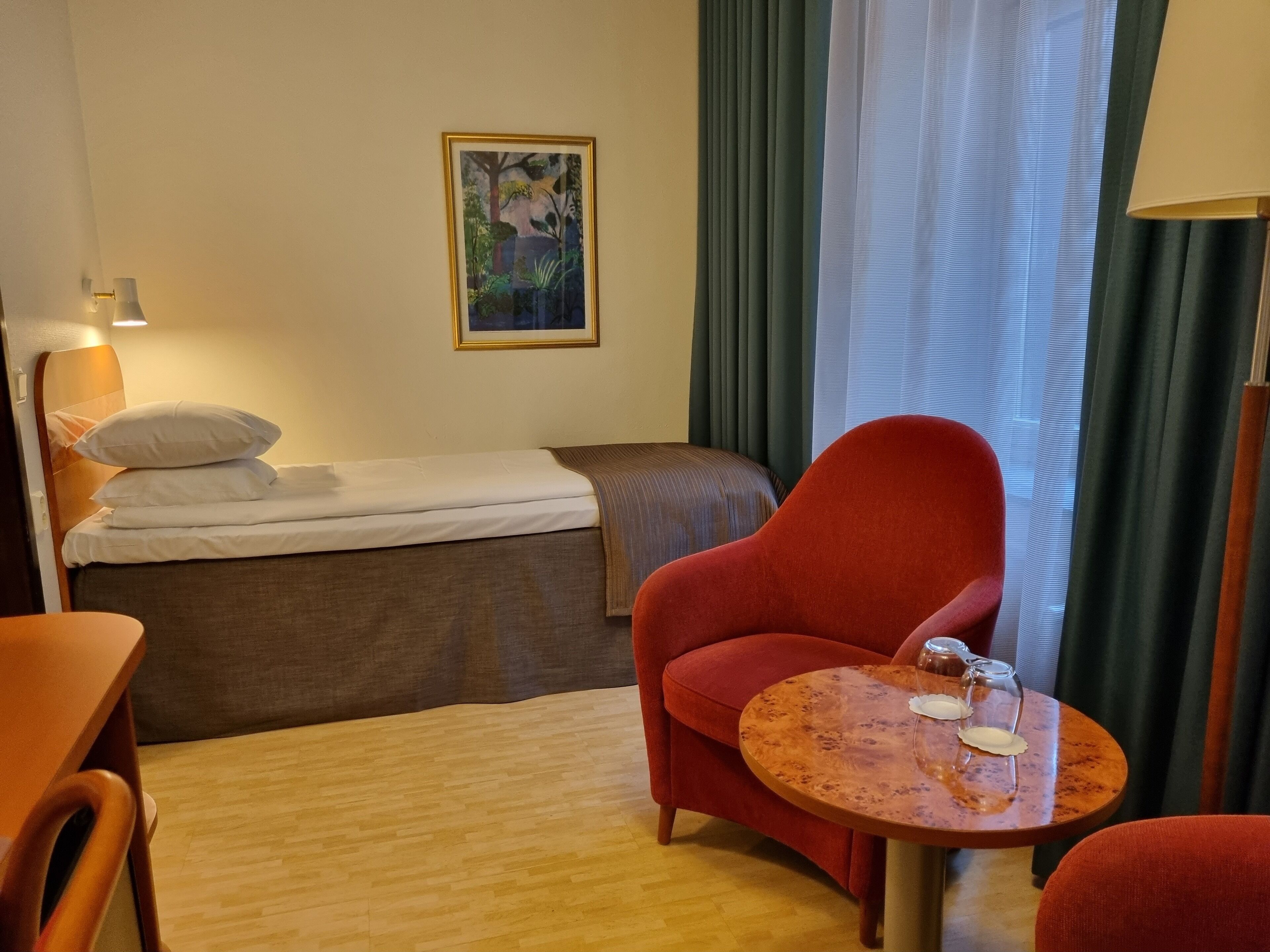 A hotel room with a bed, a red armchair, a small wooden table with a glass, and a painting on the wall.