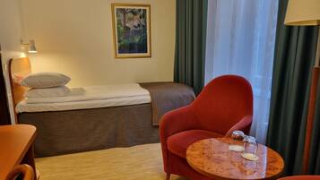 A hotel room with a bed, a red armchair, a small wooden table with a glass, and a painting on the wall.