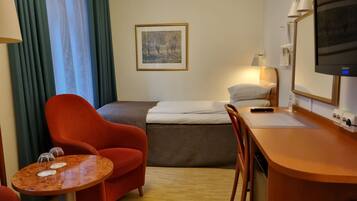 Et hotelværelse med en seng, et skrivebord med lampe, en stol, et lille bord med glas og et fjernsyn.