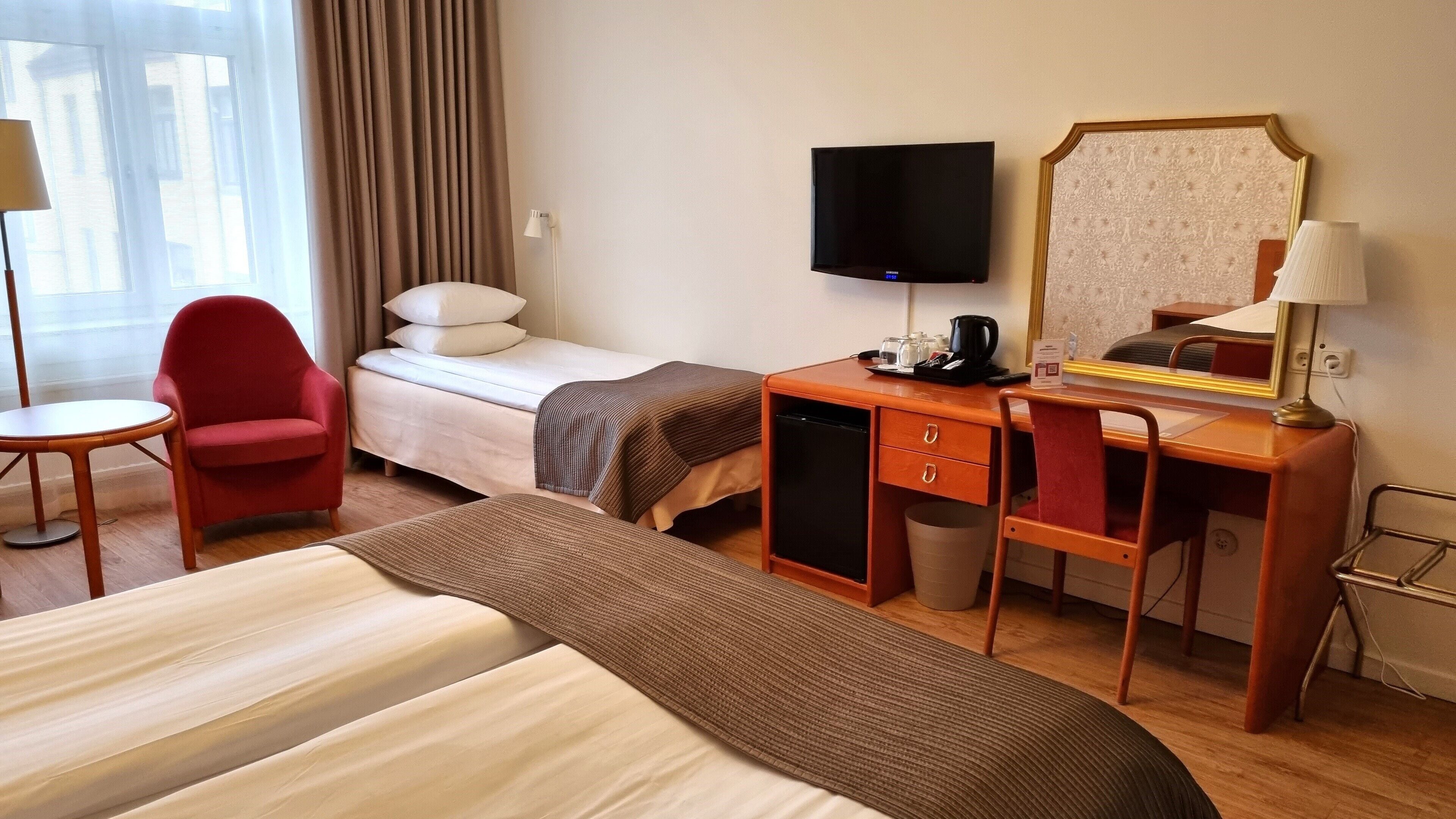 Habitación de hotel con cama, escritorio, silla, televisor y ventana con cortinas.