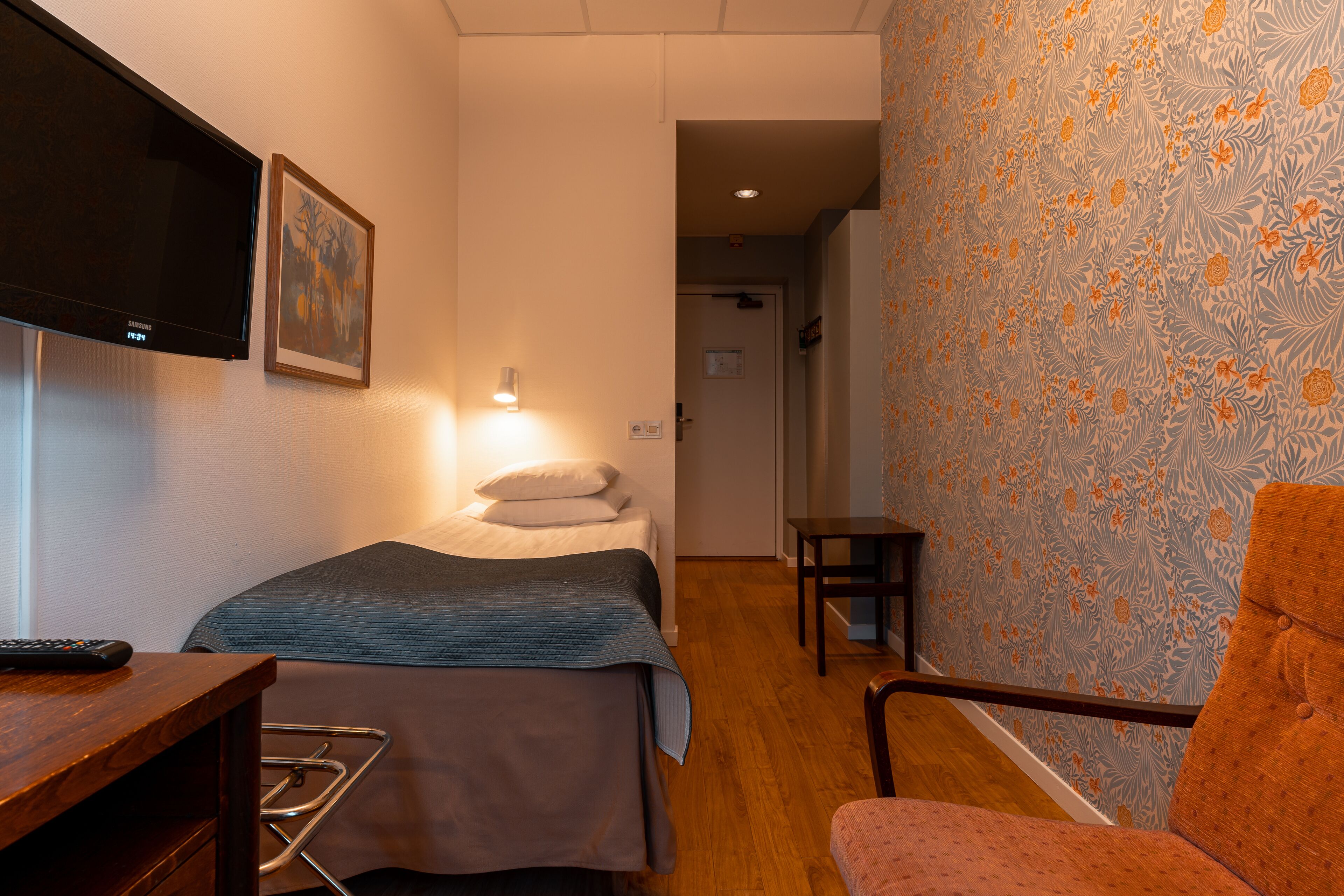 A hotel room with a bed, a TV mounted on the wall, a small table, a chair, and a nightstand.
