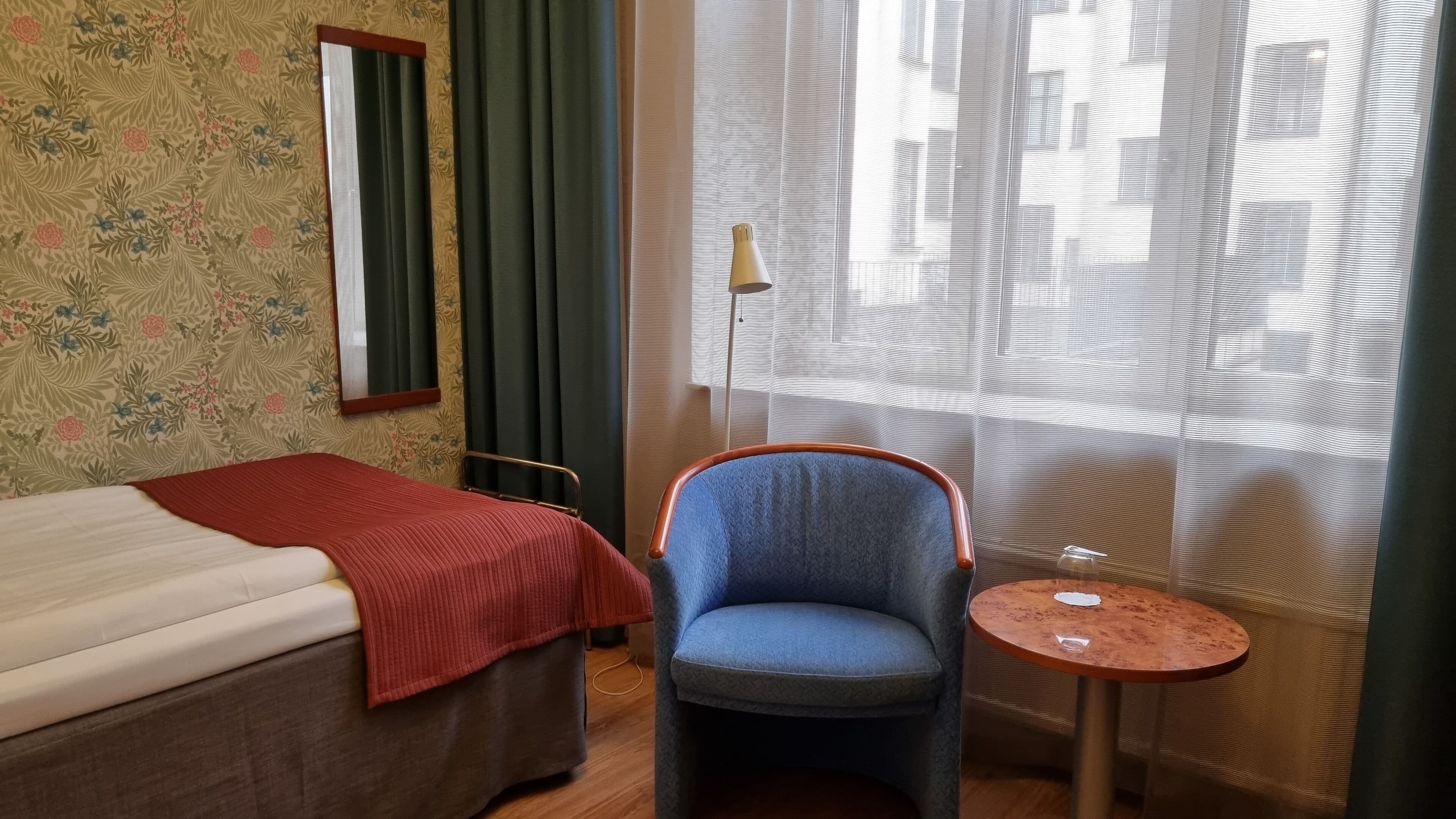 A hotel room with a bed, a chair, a small table, and a window with curtains.
