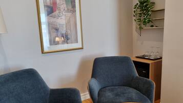 Una habitación con dos sillones grises, una mesa de madera, una obra de arte enmarco, una planta y una estantería con copas de vino.