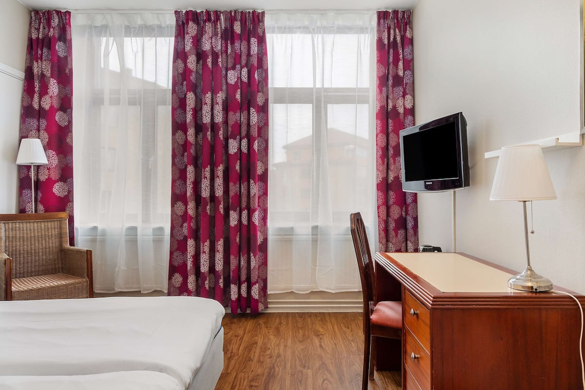 Habitación de hotel con cama, escritorio, silla, televisor y ventana con cortinas.
