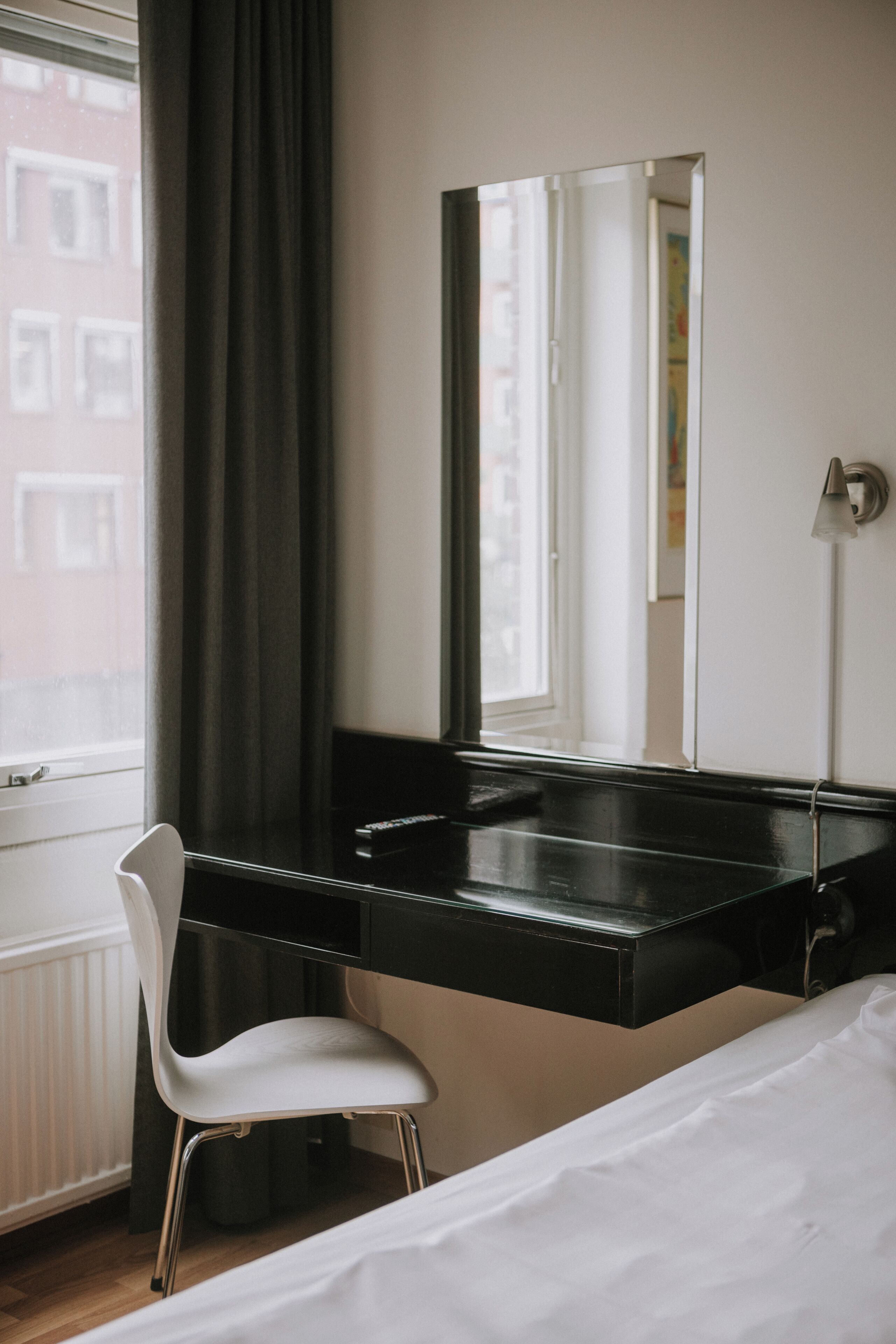 Habitación de hotel con un escritorio negro, una silla blanca, una cama, un espejo grande y una ventana con cortinas.