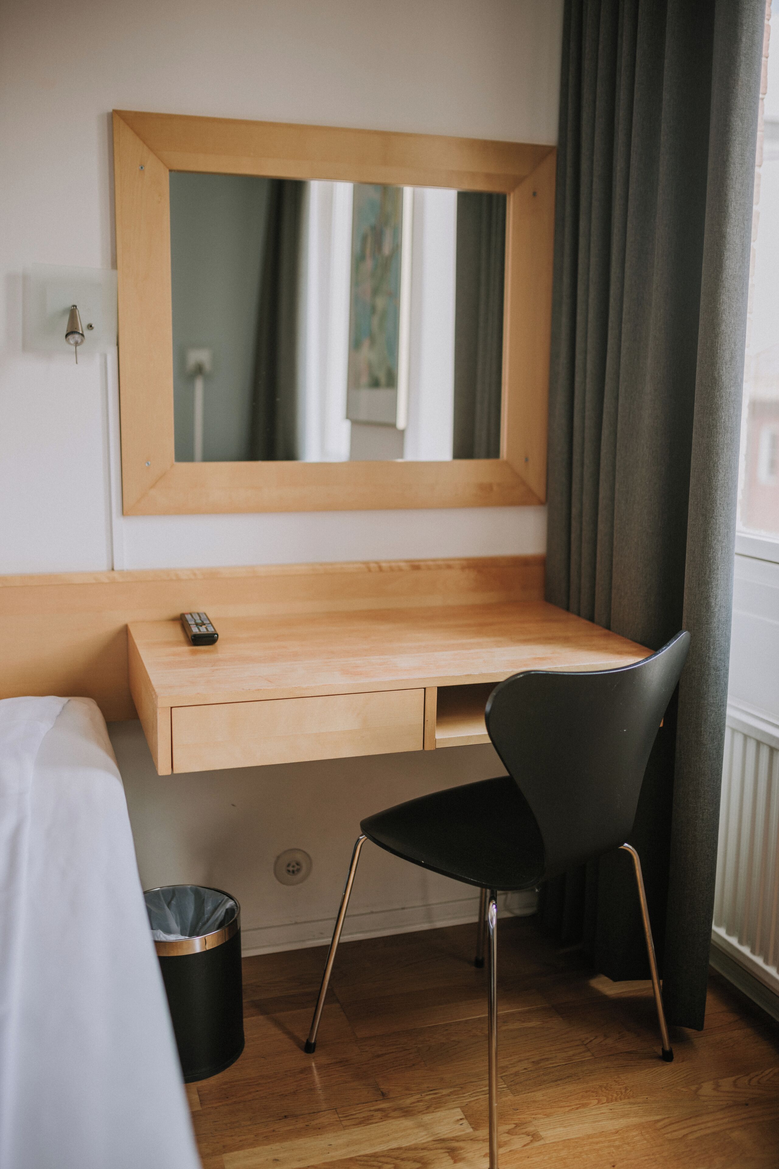 Habitación de hotel con un escritorio de madera, una silla negra, un espejo y una ventana con cortinas.