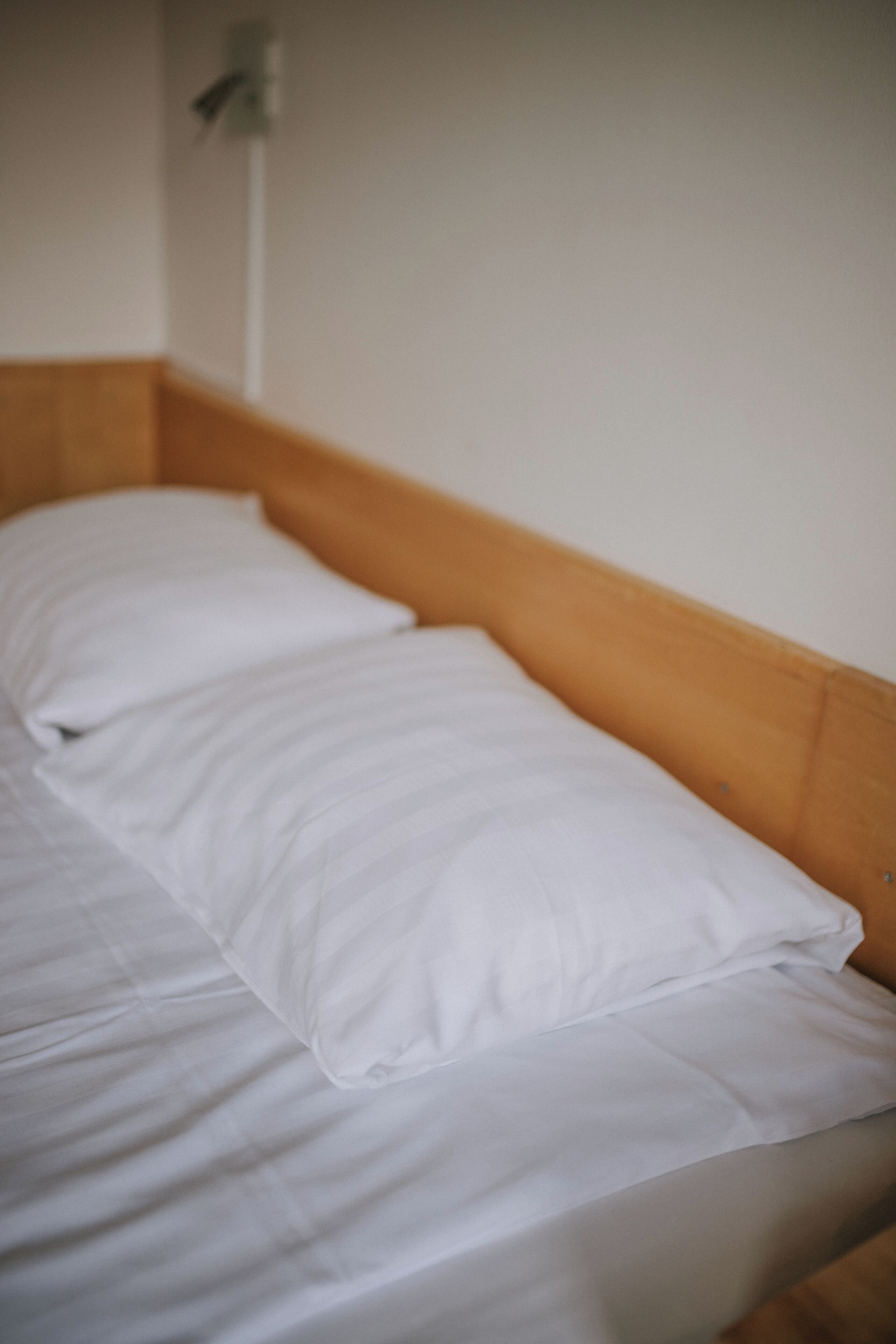 Una cama individual con sábanas blancas y cabecera de madera en una habitación con paredes lisas.