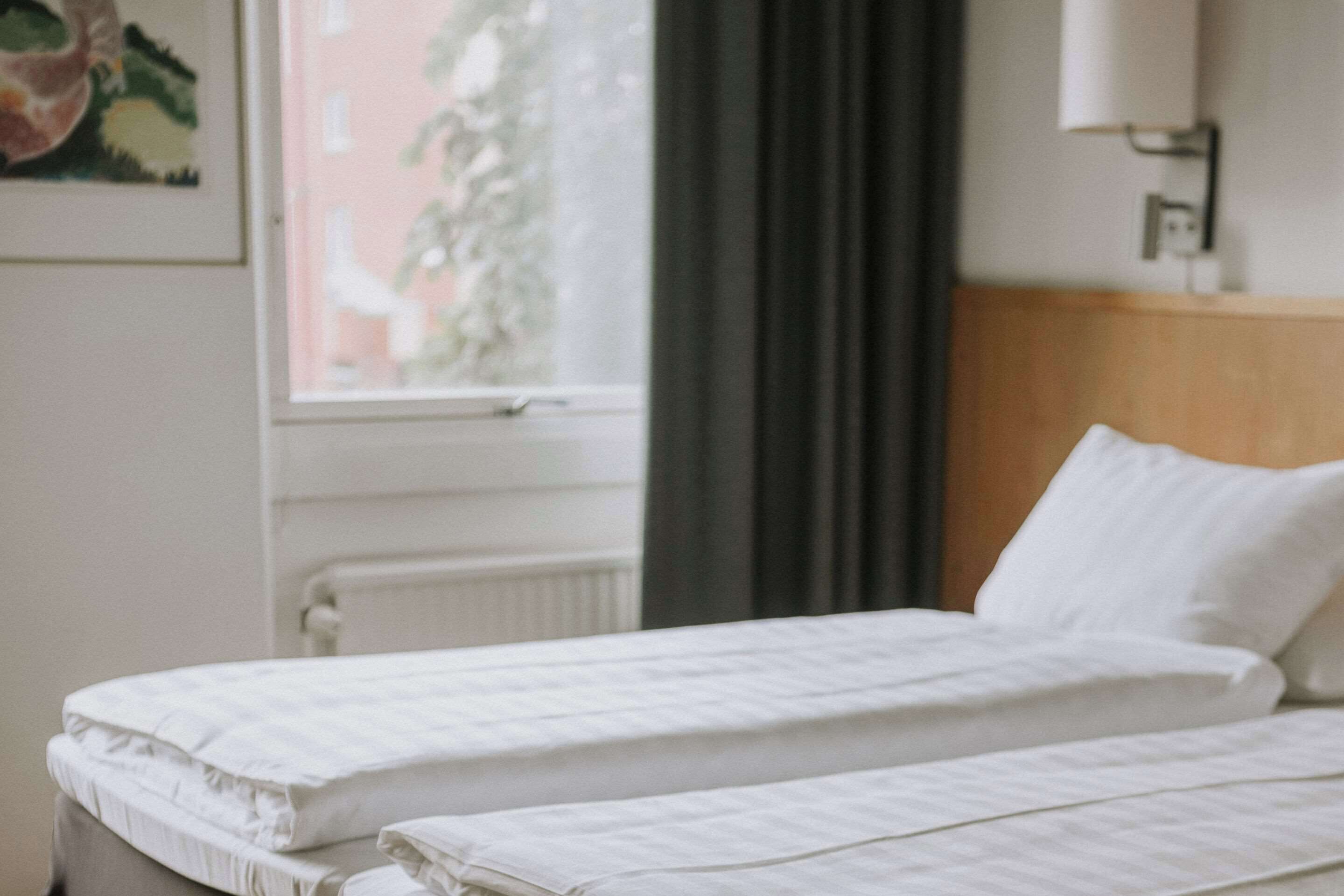 A hotel room with two single beds, a window with a view of a building and trees, and a wall-mounted light fixture.