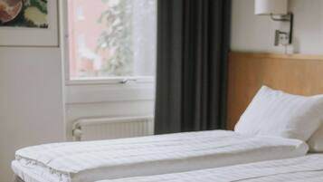 A hotel room with two single beds, a window with a view of a building and trees, and a wall-mounted light fixture.