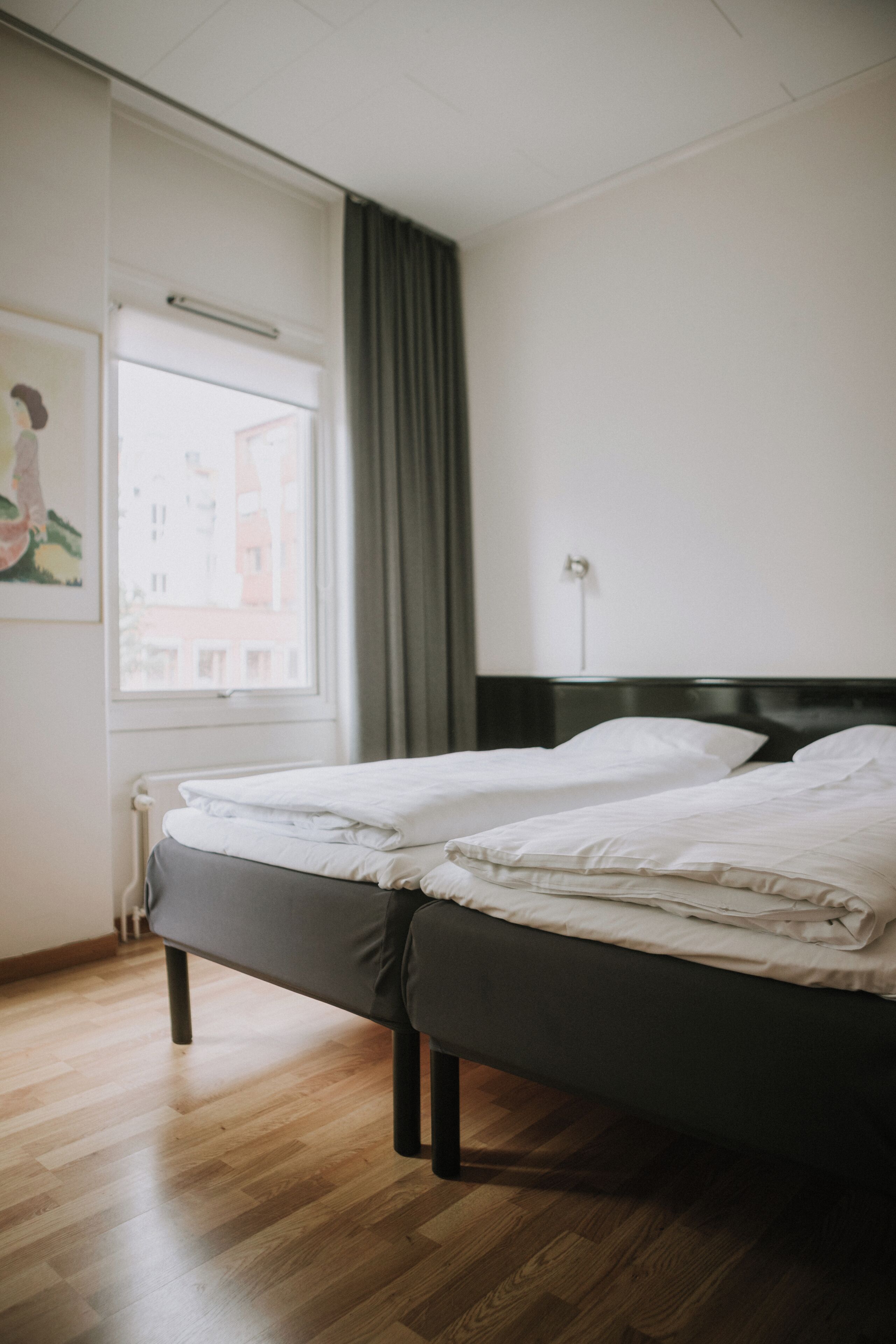A hotel room with two beds, a window with curtains, a radiator, and a wall-mounted power outlet.