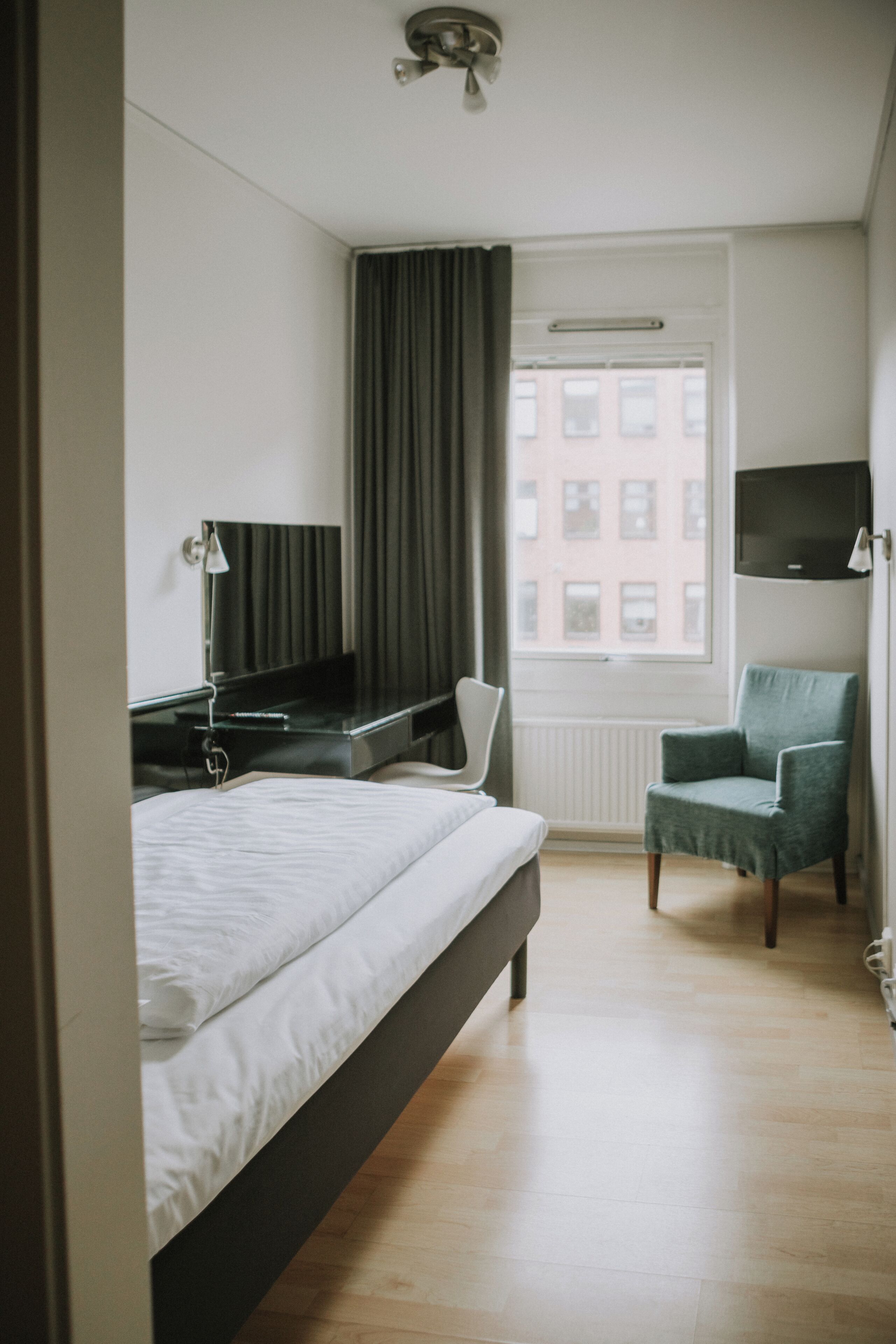 Habitación de hotel con cama, escritorio con computadora, silla, televisor y ventana con cortinas.