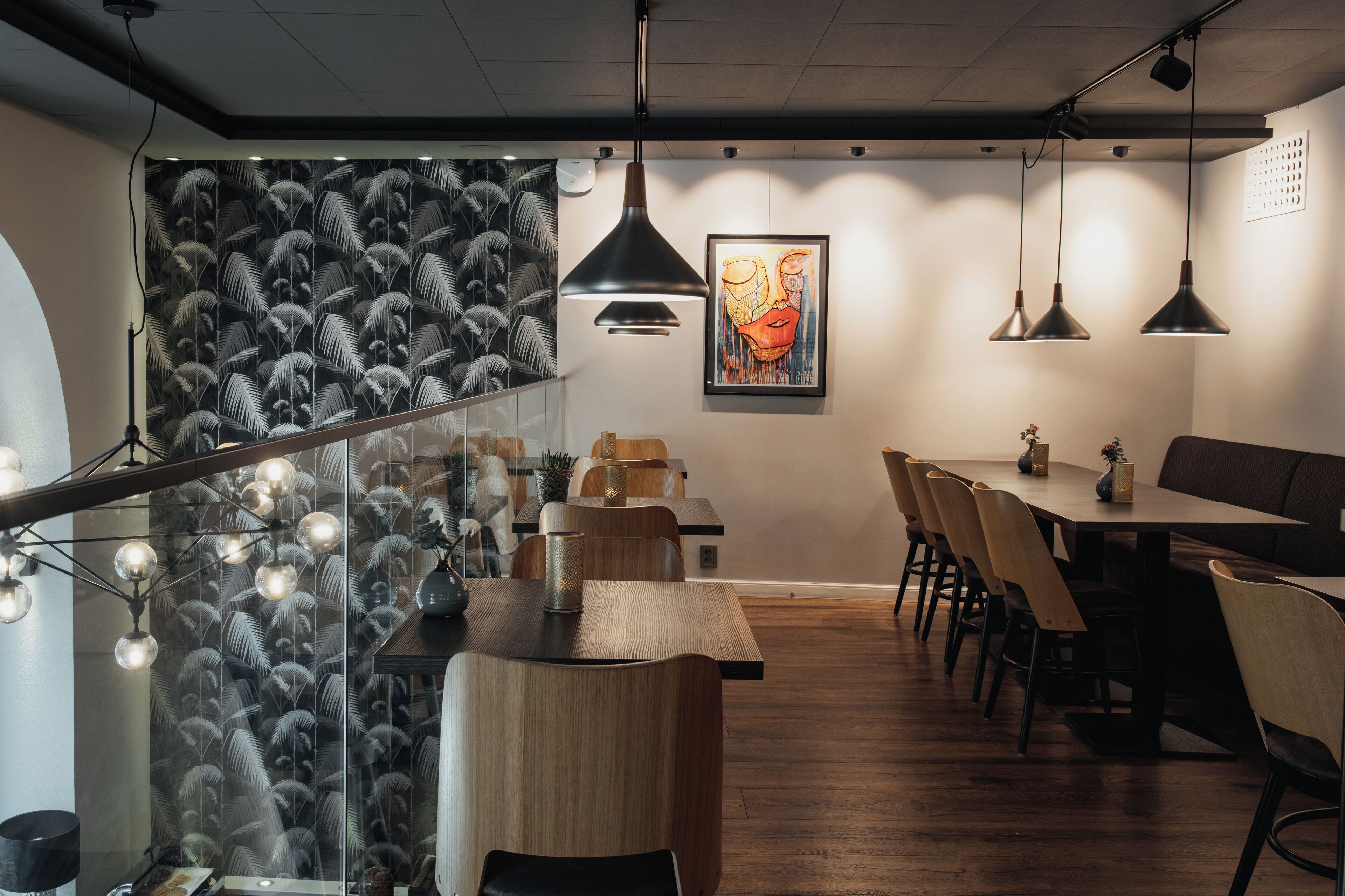 Zona pranzo moderna con tavoli e sedie in legno, illuminazione a sospensione e parete decorata con motivo a piume.