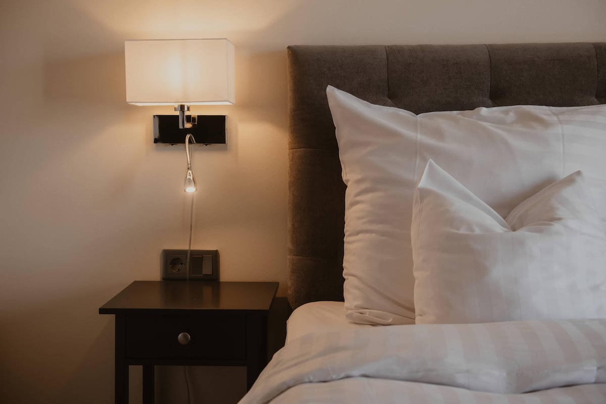 A neatly made bed with white linens, a bedside lamp, and a small nightstand with a drawer.